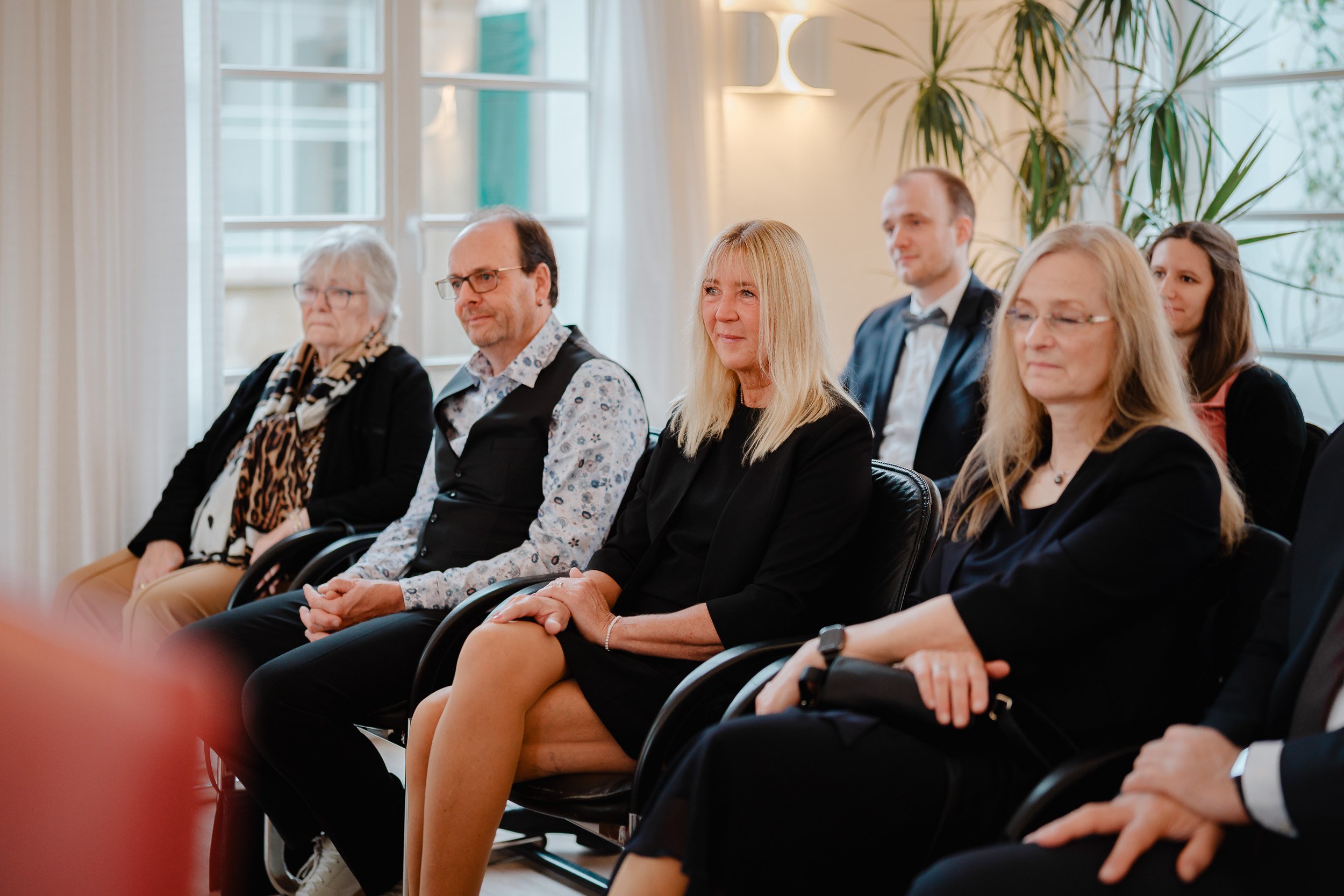Menschen sitzen in einem Raum während einer formellen Veranstaltung, gekleidet in elegante Kleidung, vor großen Fenstern mit Sicht auf das Gebäude außerhalb und einigen Pflanzen im Hintergrund.
