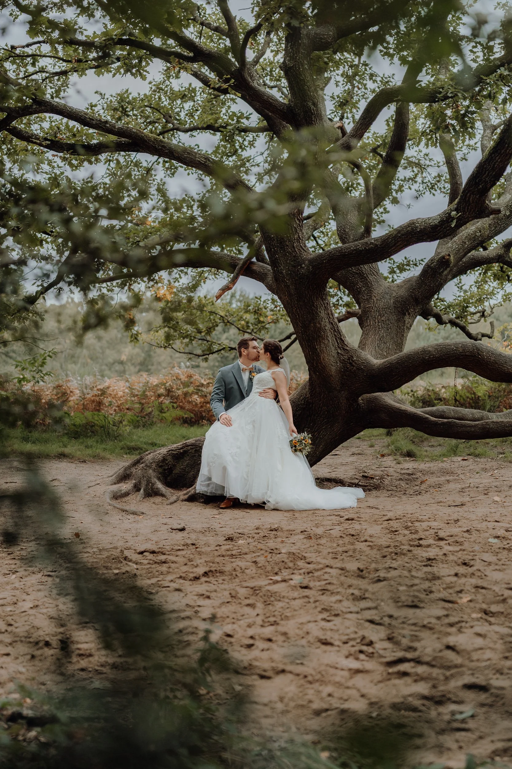 Ein Hochzeitspaar steht vor einem großen Baum in einer natürlichen Umgebung. Das Brautpaar schaut sich verliebt an, die Braut hält einen Blumenstrauß, der Bräutigam trägt einen grauen Anzug.