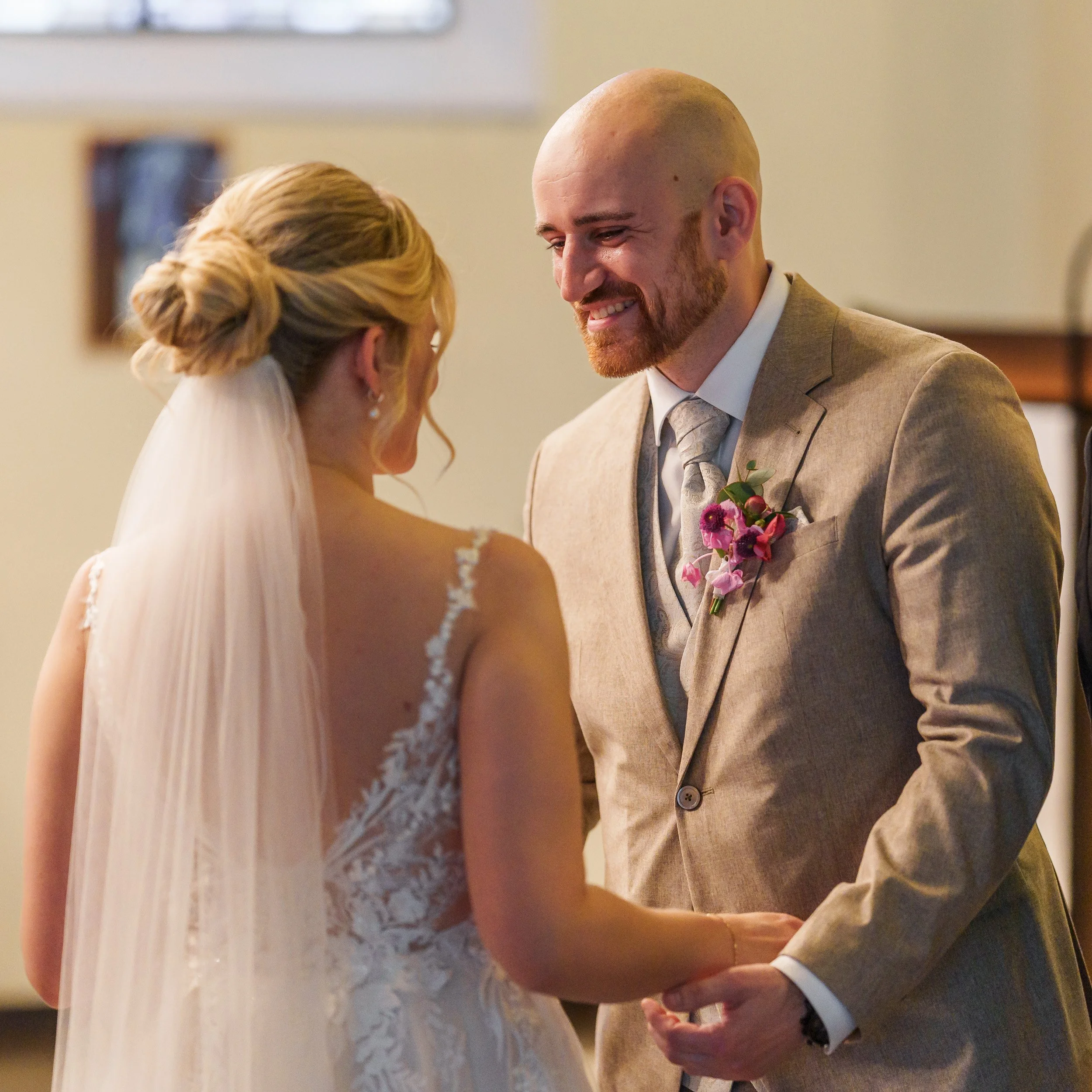 Ein Brautpaar bei der Trauung, er hält ihre Hände, sie trägt ein Brautkleid mit Spitzenverzierungen, er trägt einen hellbraunen Anzug mit einer Blume im Revers.