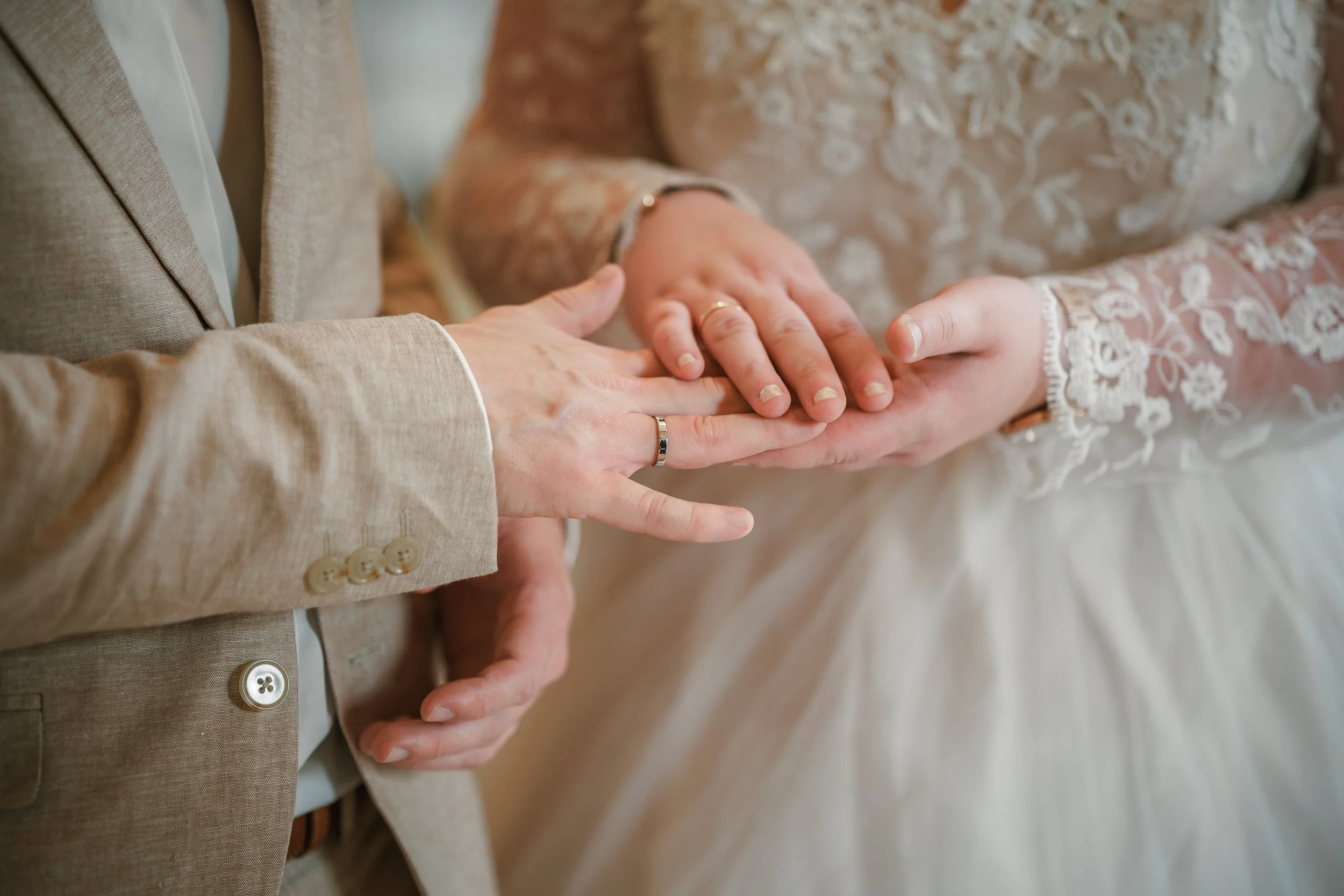 Nahaufnahme von drei Händen, die sich berühren. Eine Hand eines Paares hält die Hand eines Babys, alle mit Ringen. Das Paar trägt elegante, helle Kleidung, eine im Jackett, die andere in einem Spitzenkleid.