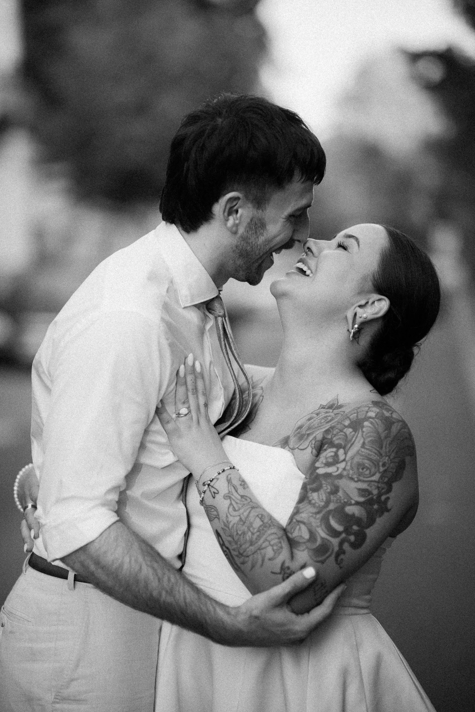 Ein lachendes Paar, vermutlich bei einer Hochzeit, umarmt sich im Freien. Der Mann trägt ein Hemd und die Frau hat Tattoos an den Armen und trägt ein Kleid.
