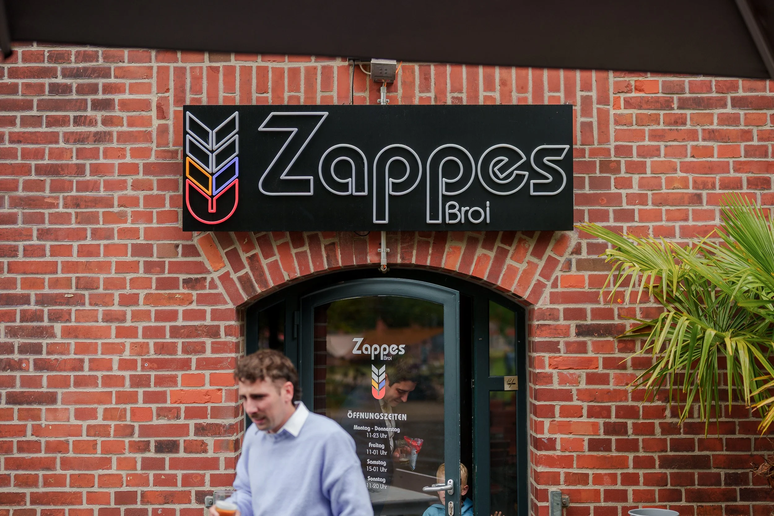 Ein Laden mit einem schwarzen Neon-Logo-Schilder über der Tür, das den Namen 'Zappes Broi' zeigt, an einer roten Backsteinfassade, mit Menschen im Vordergrund, einer davon mit einem Getränk in der Hand.