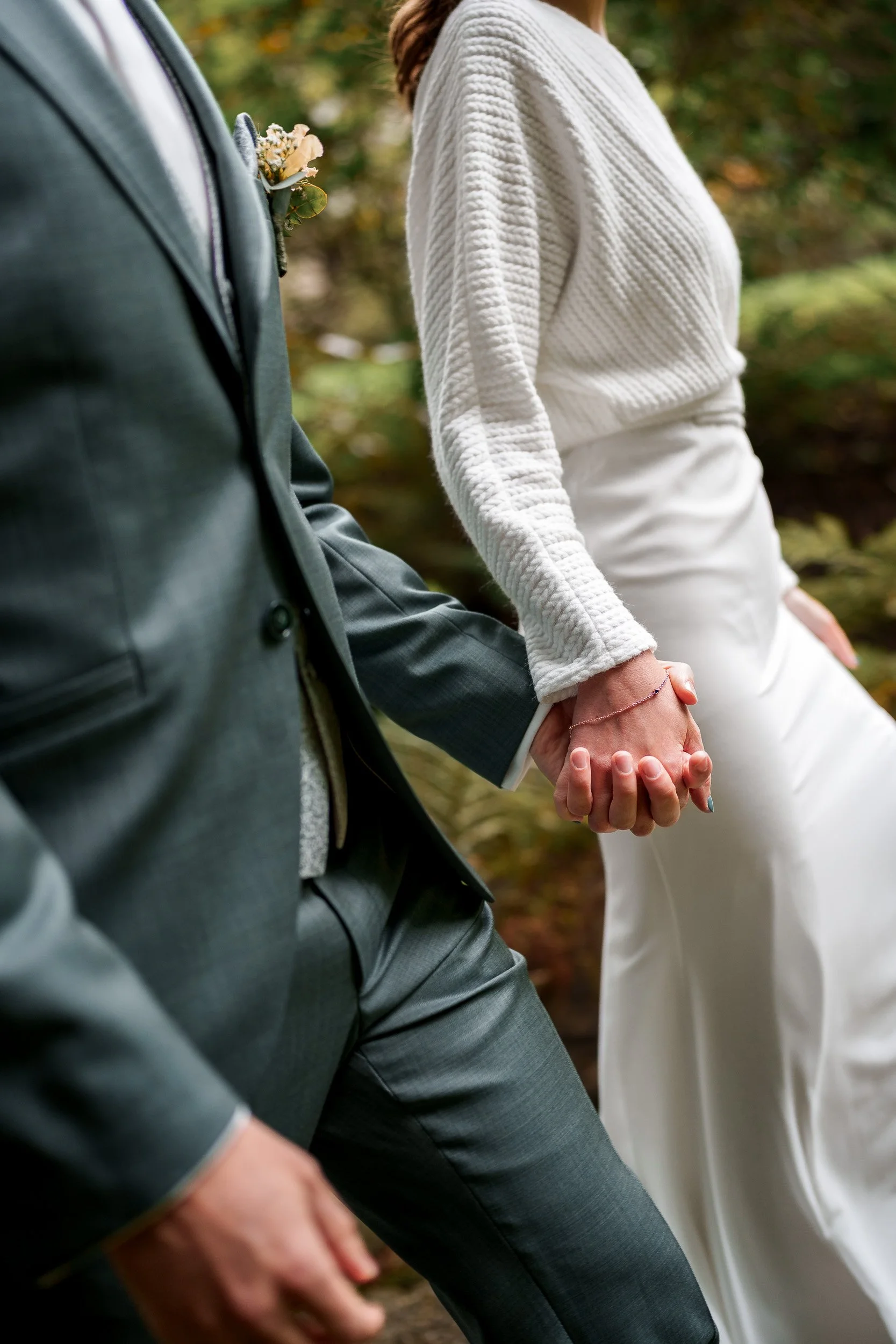 Ein Hochzeitsfoto von einem Mann in einem dunklen Anzug und einer Frau in einem weißen Outfit, die sich gegenseitig die Hände halten.