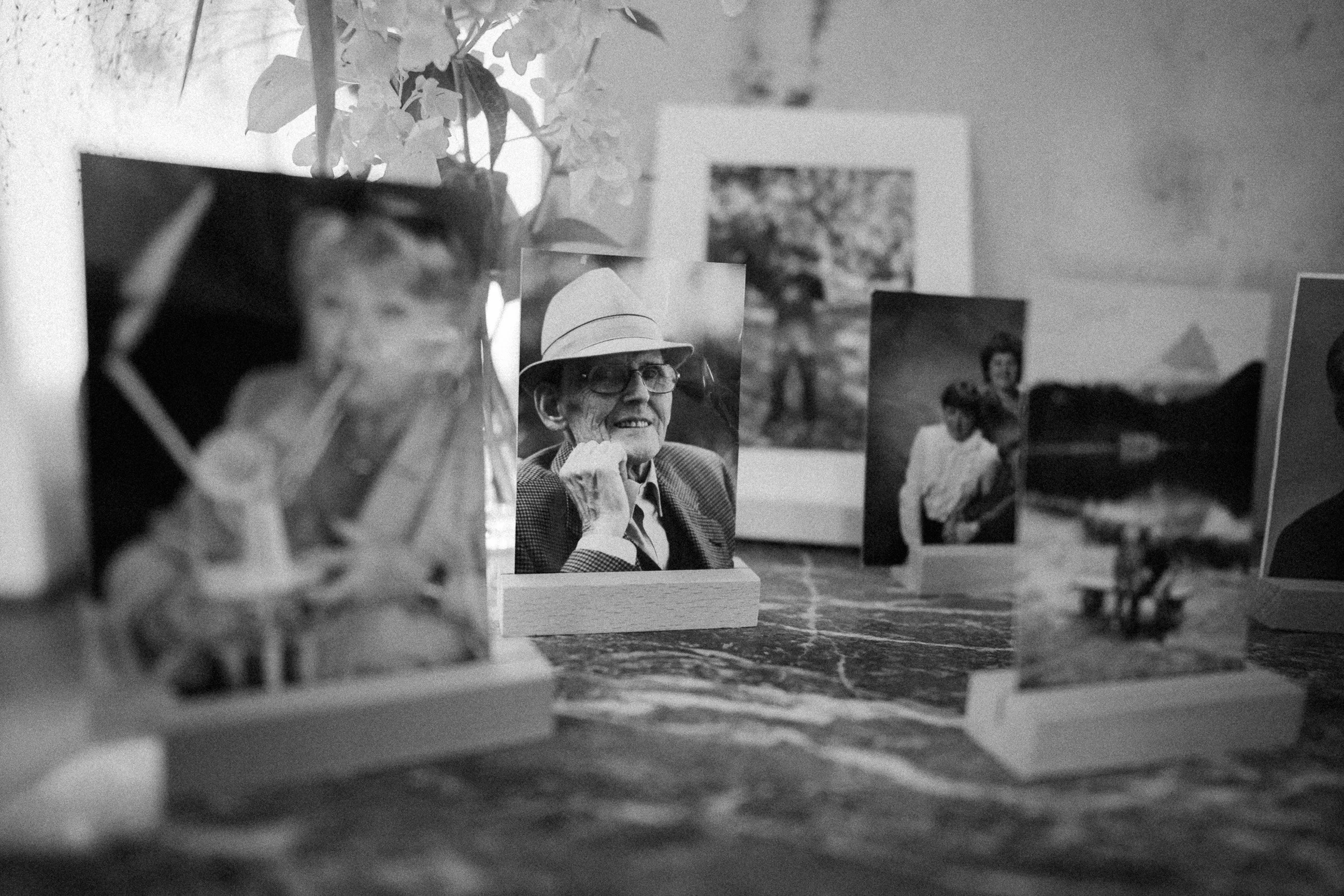 Schwarz-weiß-Fotografie einer Sammlung von Fotos auf einem Tisch, darunter ein älterer Mann mit Brille und Hut, involviert in ein Erinnerungs- oder Gedenkprojekt.