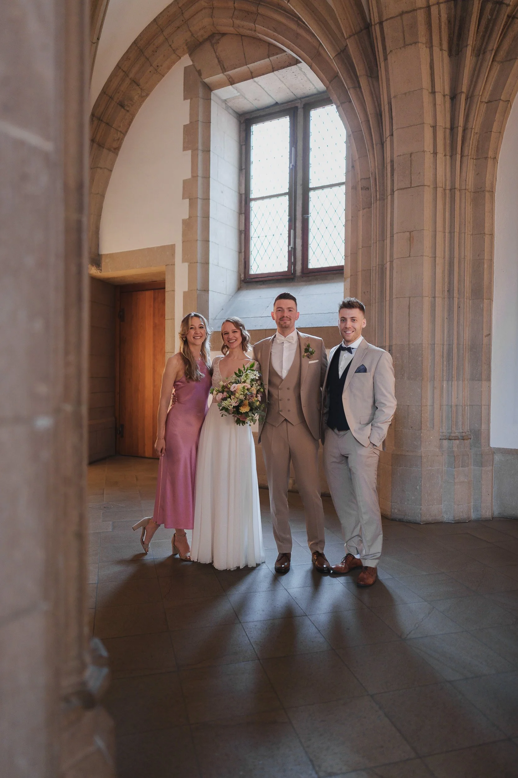 Vier Menschen in formeller Kleidung, zwei Frauen und zwei Männer, stehen im Inneren eines historischen Gebäudes mit Steinbögen und großem Fenster im Hintergrund, bei einer Hochzeit.