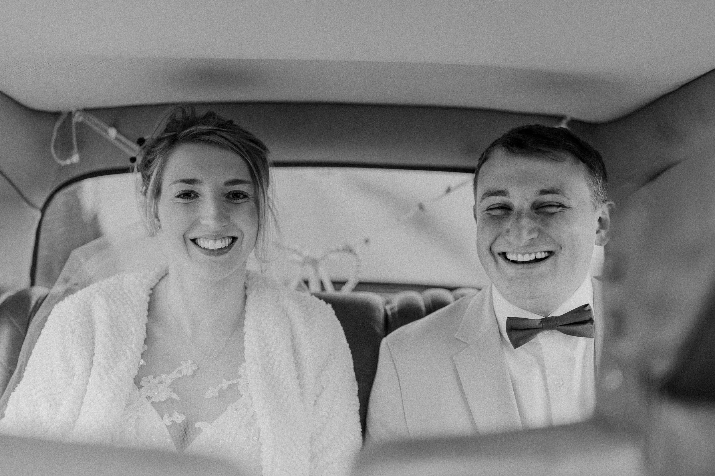Ein glückliches Brautpaar sitzt in einem vintage Auto, beide lachen und tragen formelle Hochzeitskleidung, die Frau trägt ein Brautkleid mit Spitze, der Mann einen Anzug mit Fliege, schwarz-weiß Fotografie.