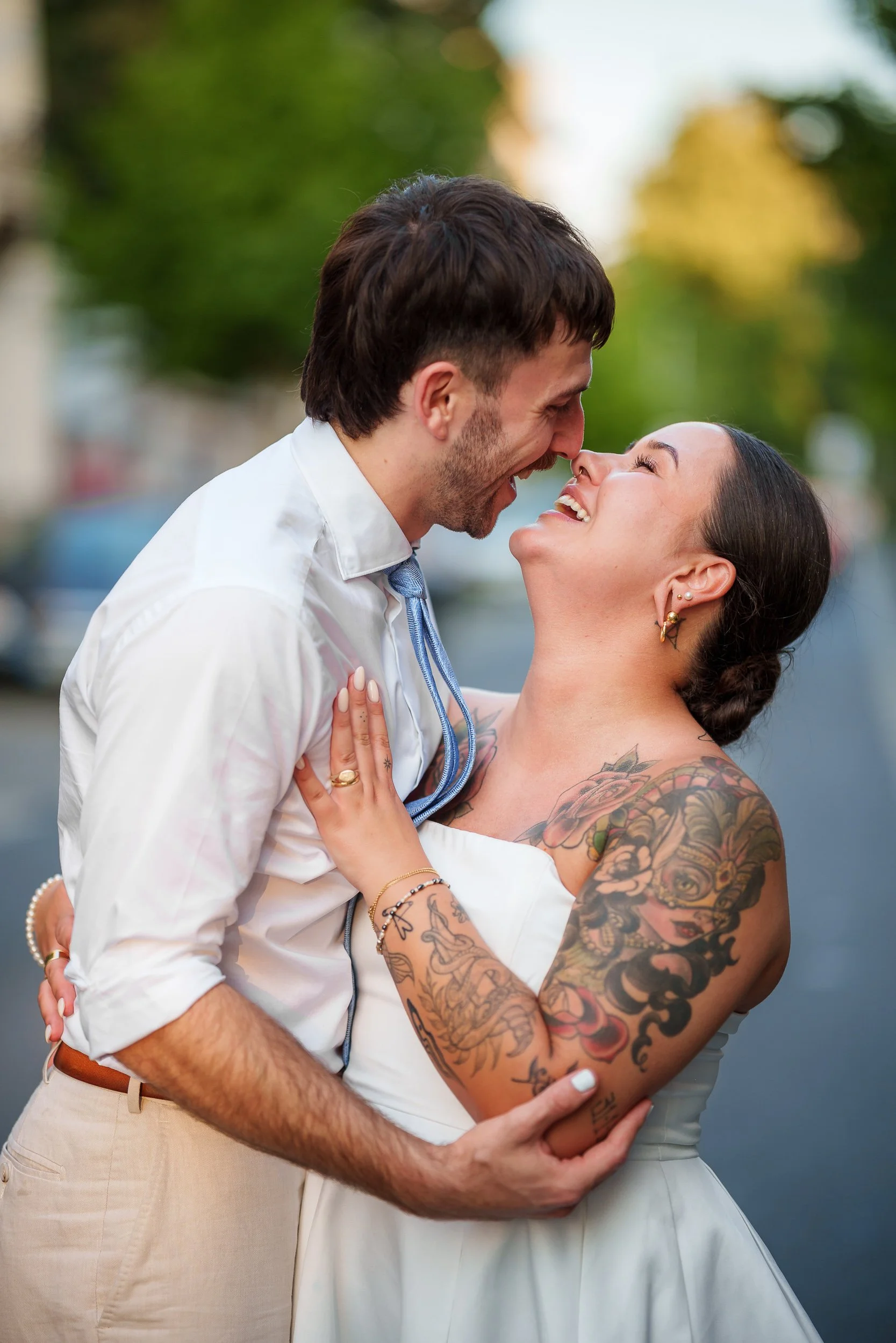 Ein glücklich schauendes Paar, das sich umarmt und sich liebevoll gegenübersieht, draußen bei Sonnenuntergang, mit sichtbaren Tattoos auf den Armen der Frau.