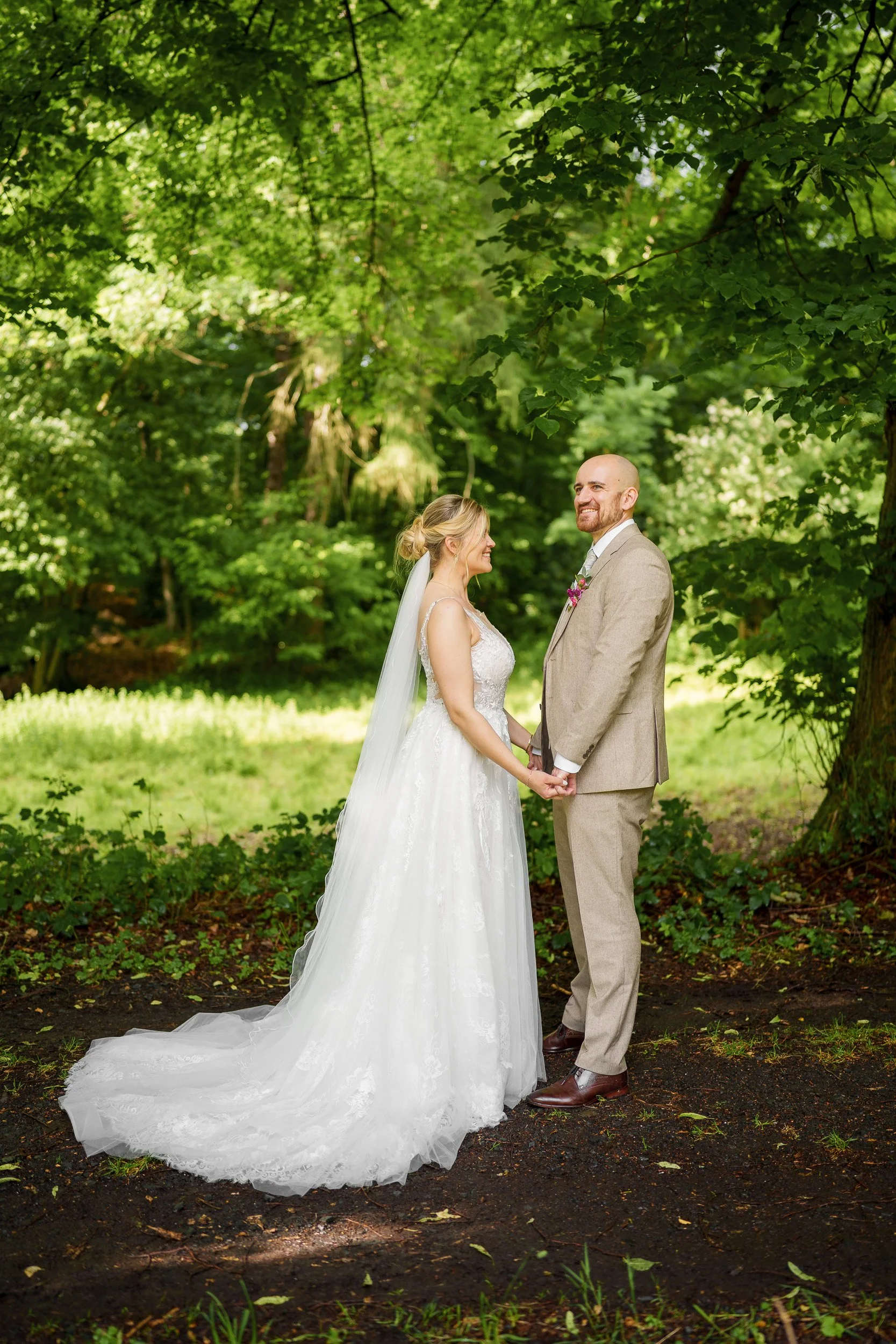 Braut und Bräutigam stehen sich im Wald gegenüber, halten Händchen, lächeln sich an, umgeben von grünen Bäumen an einem sonnigen Tag.