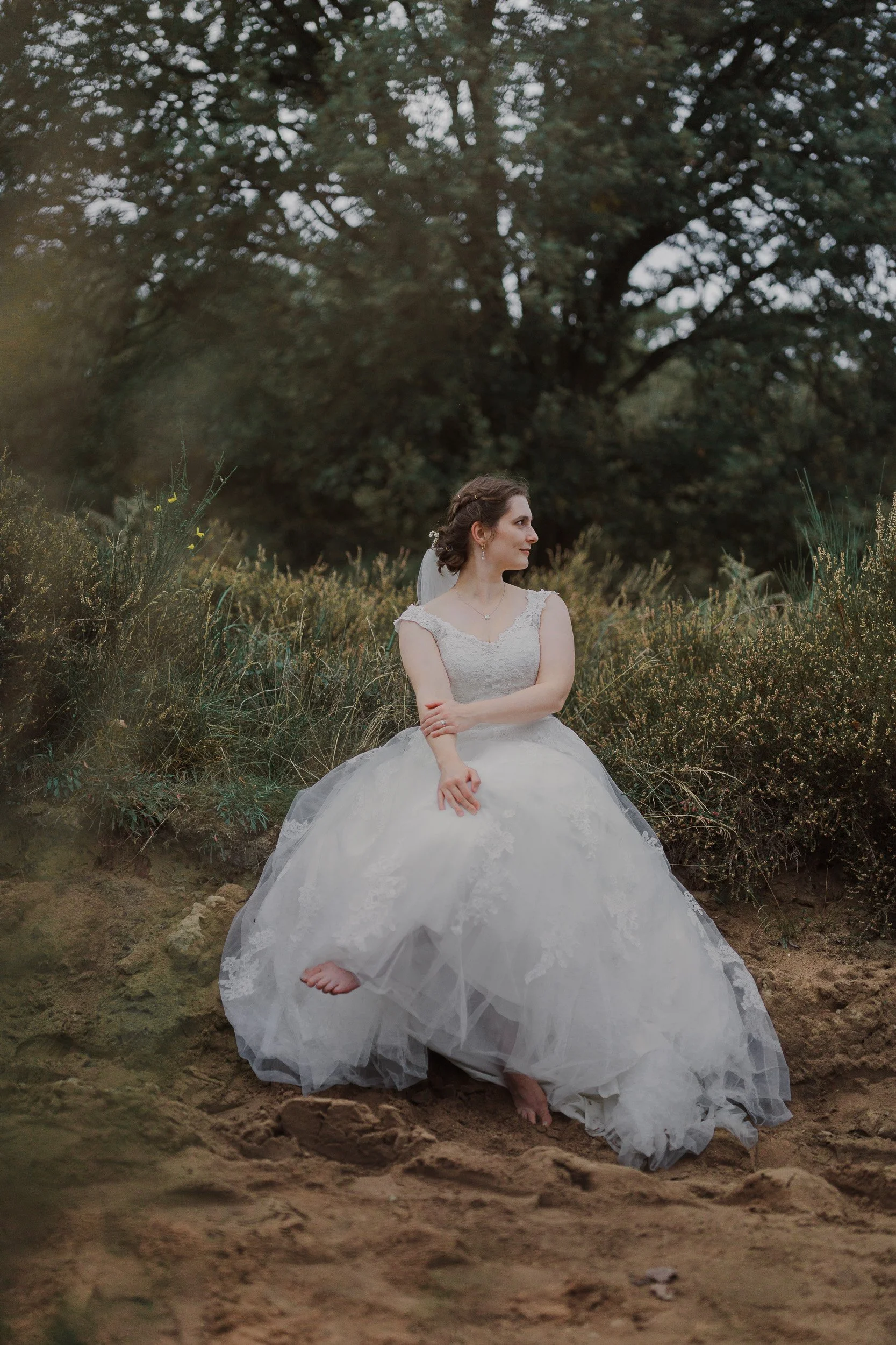 Eine Frau in einem weißen Hochzeitskleid sitzt barfuß im Sand vor einer Buschwiese, im Hintergrund Bäume, bei Sonnenuntergang.