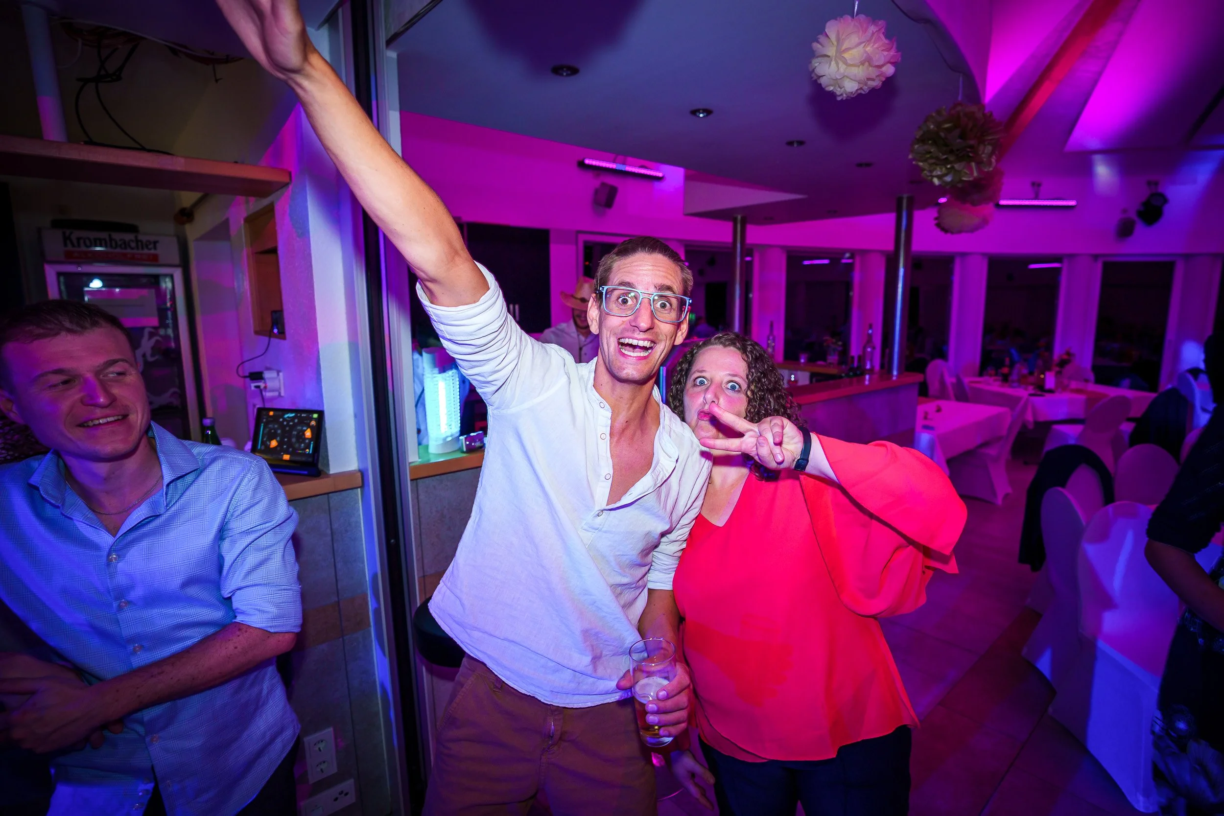 Menschen feiern in einer festlich dekorierten Location mit buntem Licht, lachen und posieren für die Kamera, einer mit Brille und einem Glas in der Hand, eine Frau macht ein Peace-Zeichen.