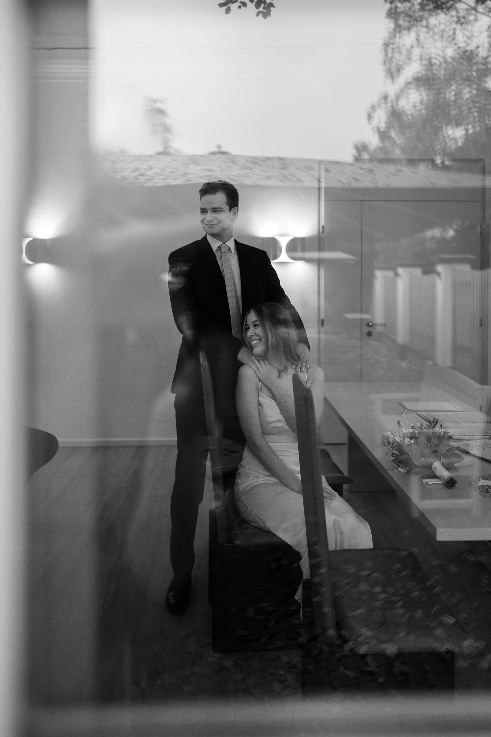 Ein Mann in Anzug steht neben einer sitzenden Frau in einem weißen Kleid, beide lachen und spielen auf einem Raum, gesehen durch eine Fensterscheibe.