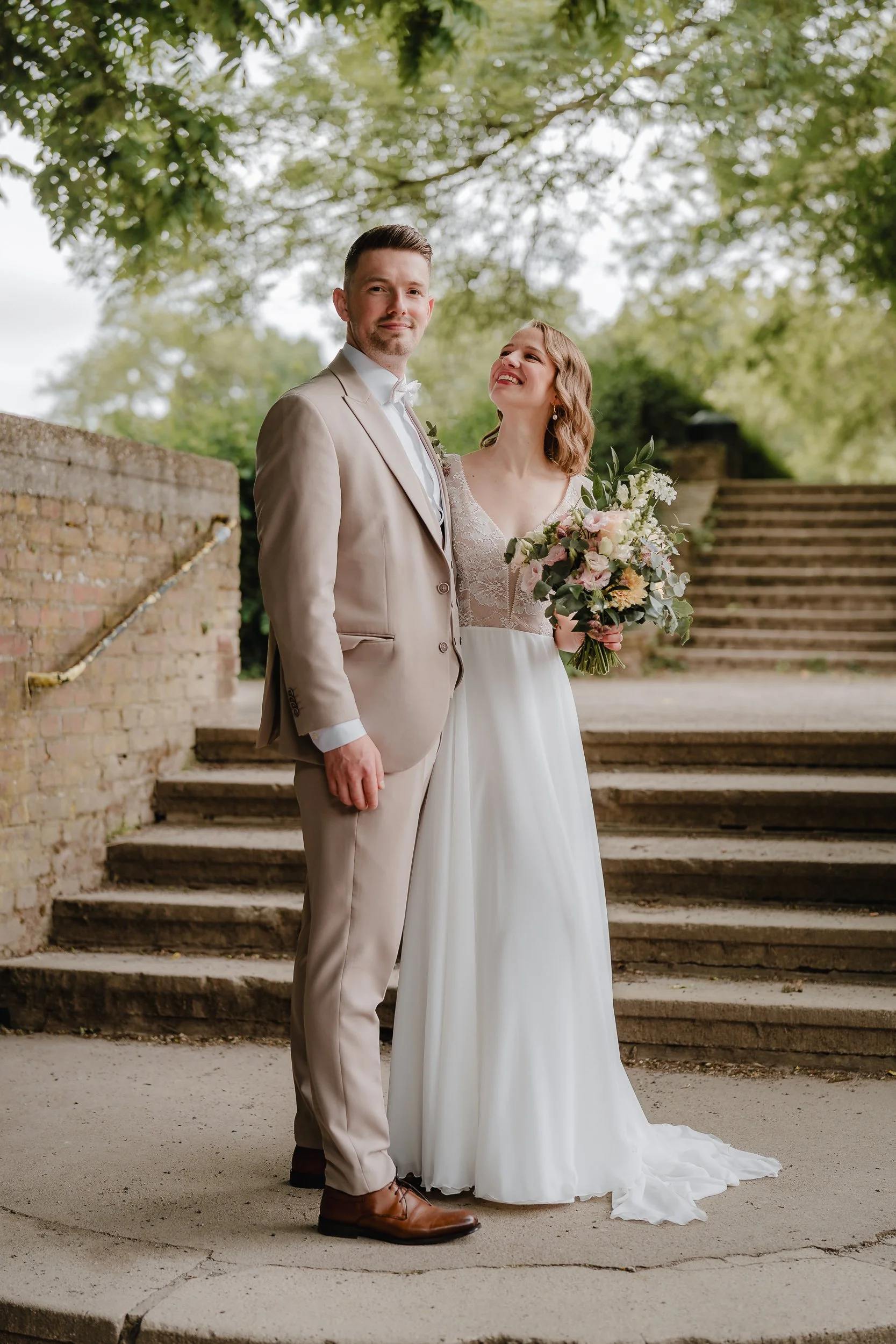 Ein frisch verheiratetes Paar steht auf einer Treppe im Freien. Die Frau hält einen Blumenstrauß und trägt ein weißes Brautkleid. Der Mann trägt einen beige Anzug. Sie sind umgeben von grünen Bäumen.