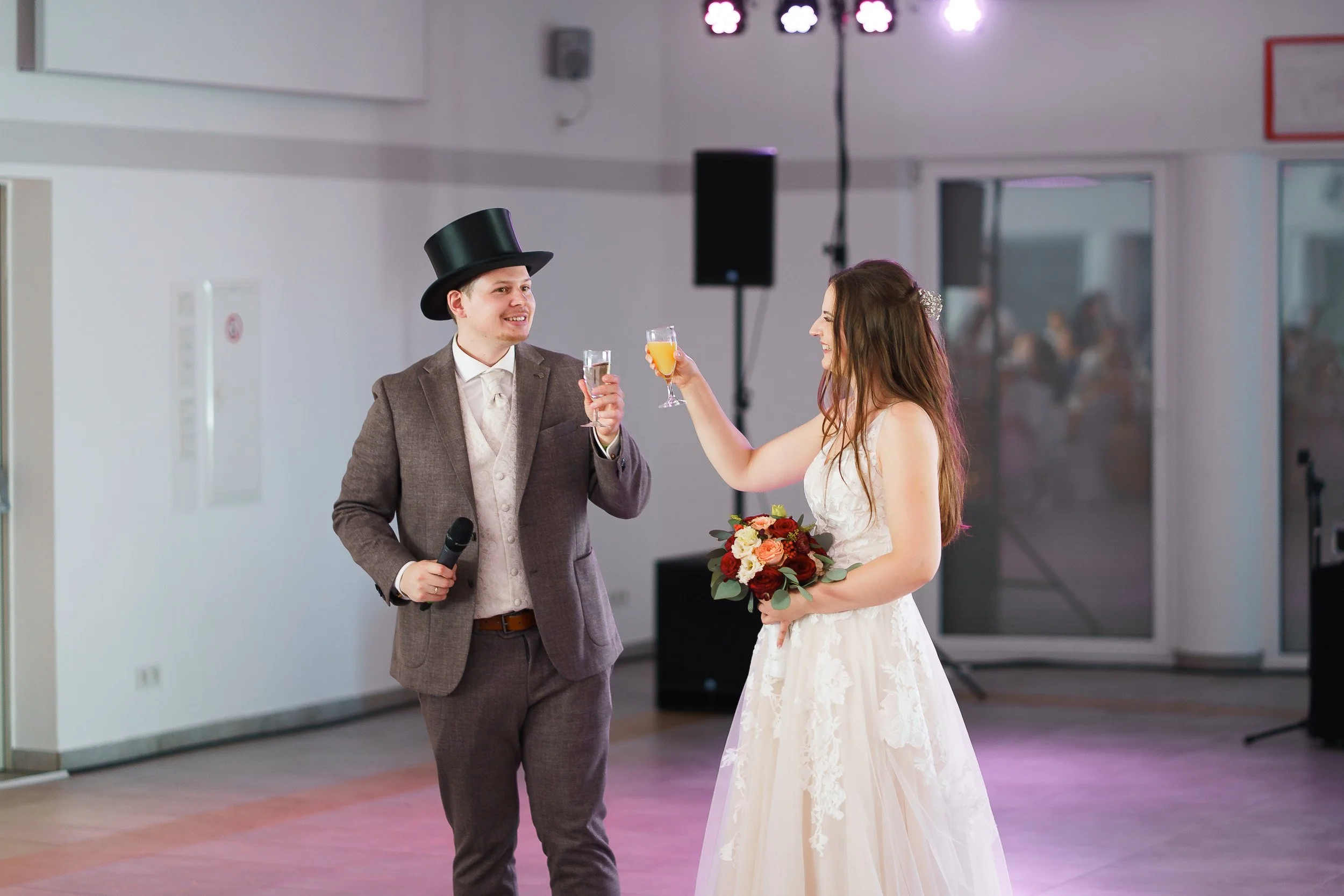Ein Brautpaar bei einer Hochzeitsfeier, das gemeinsam anstößt. Die Braut trägt ein weißes Hochzeitskleid und hält einen Blumenstrauß, während der Bräutigam einen Anzug trägt mit einem Zylinder auf dem Kopf. Im Hintergrund sind Gäste und technische Au