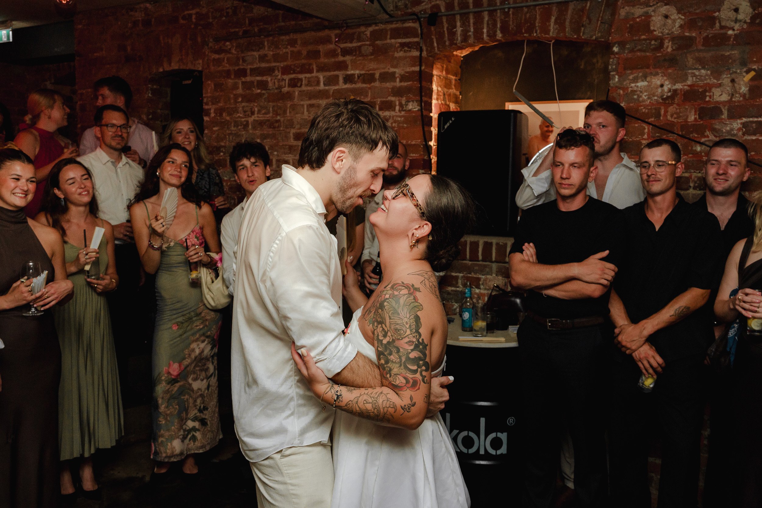 Ein junges Paar tanzt inmitten einer Gruppe von feiern den Hochzeit. Die Frau trägt ein weißes Kleid und hat Tattoos an den Armen, der Mann trägt ein weißes Hemd. Die Gäste um sie herum lachen und schauen das Paar an, während sie auf einer Party in e