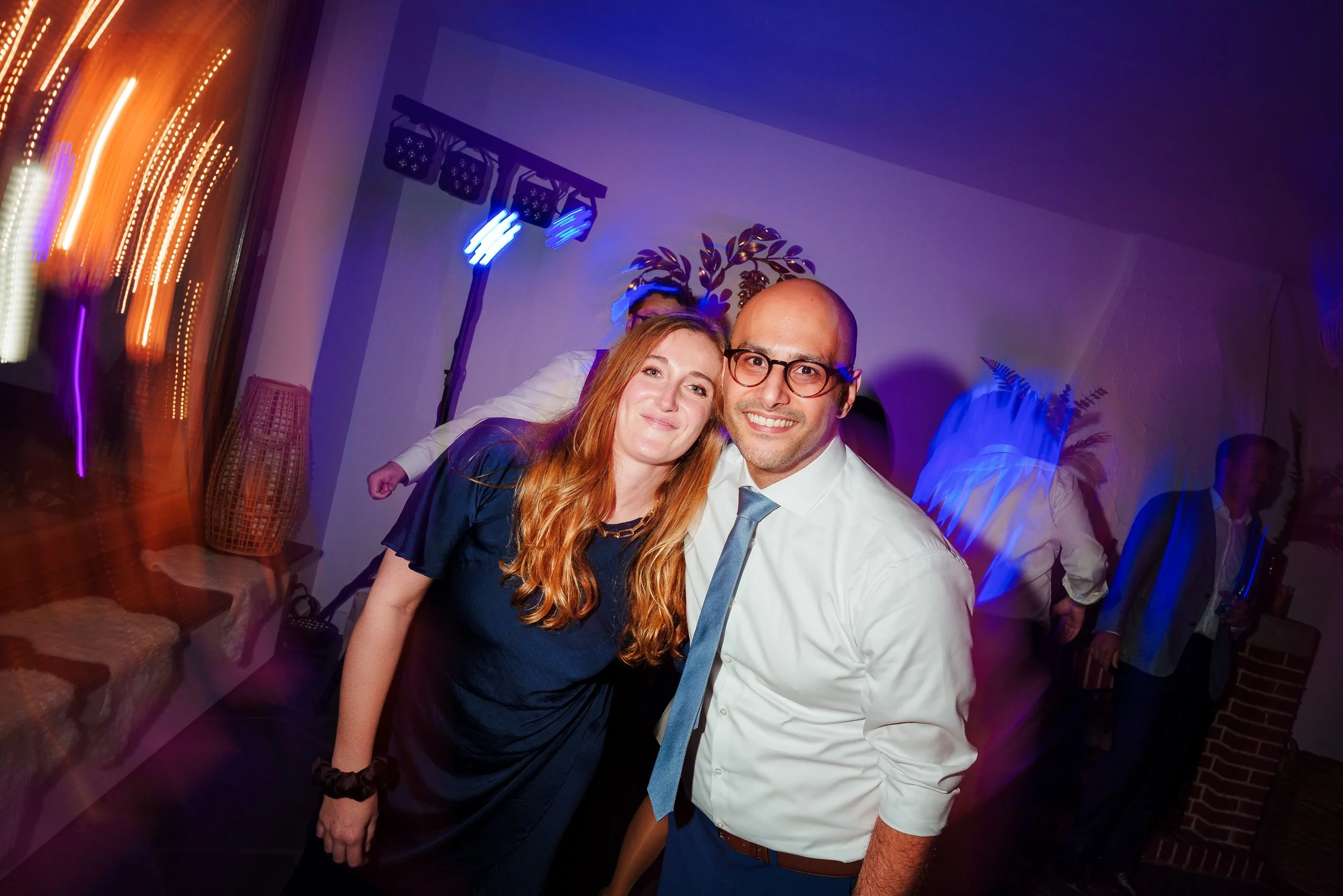 Zwei Menschen lächeln bei einer Party im Innenraum, Hintergrund mit bunten Lichtern und anderen Gästen.