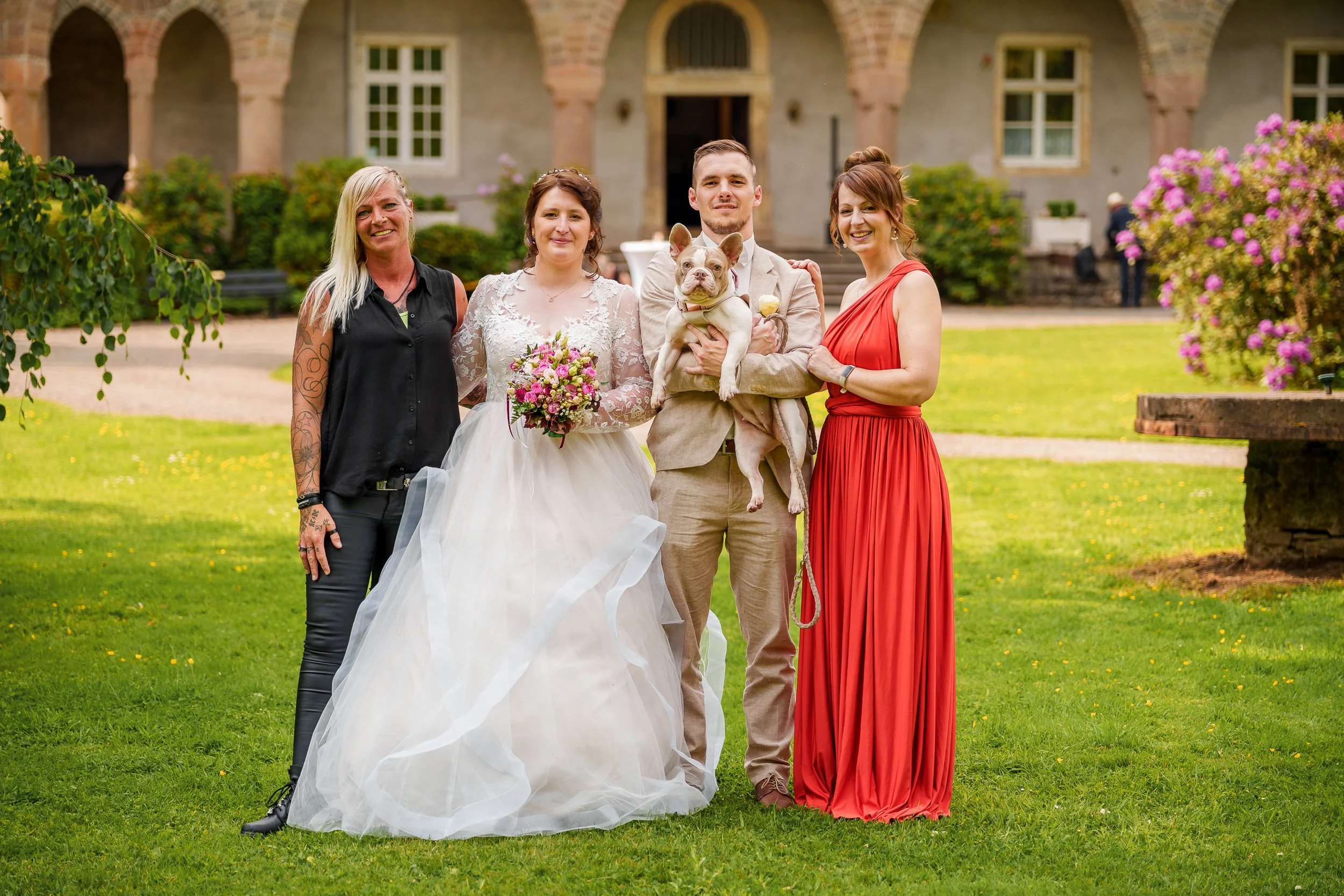 Gruppe von fünf Personen, darunter eine Braut in Hochzeitskleid mit Blumenstrauß, ein Bräutigam mit Hund, und drei Frauen in unterschiedlichen Kleidern, im Garten vor einem alten Gebäude mit Büschen und Blumen.