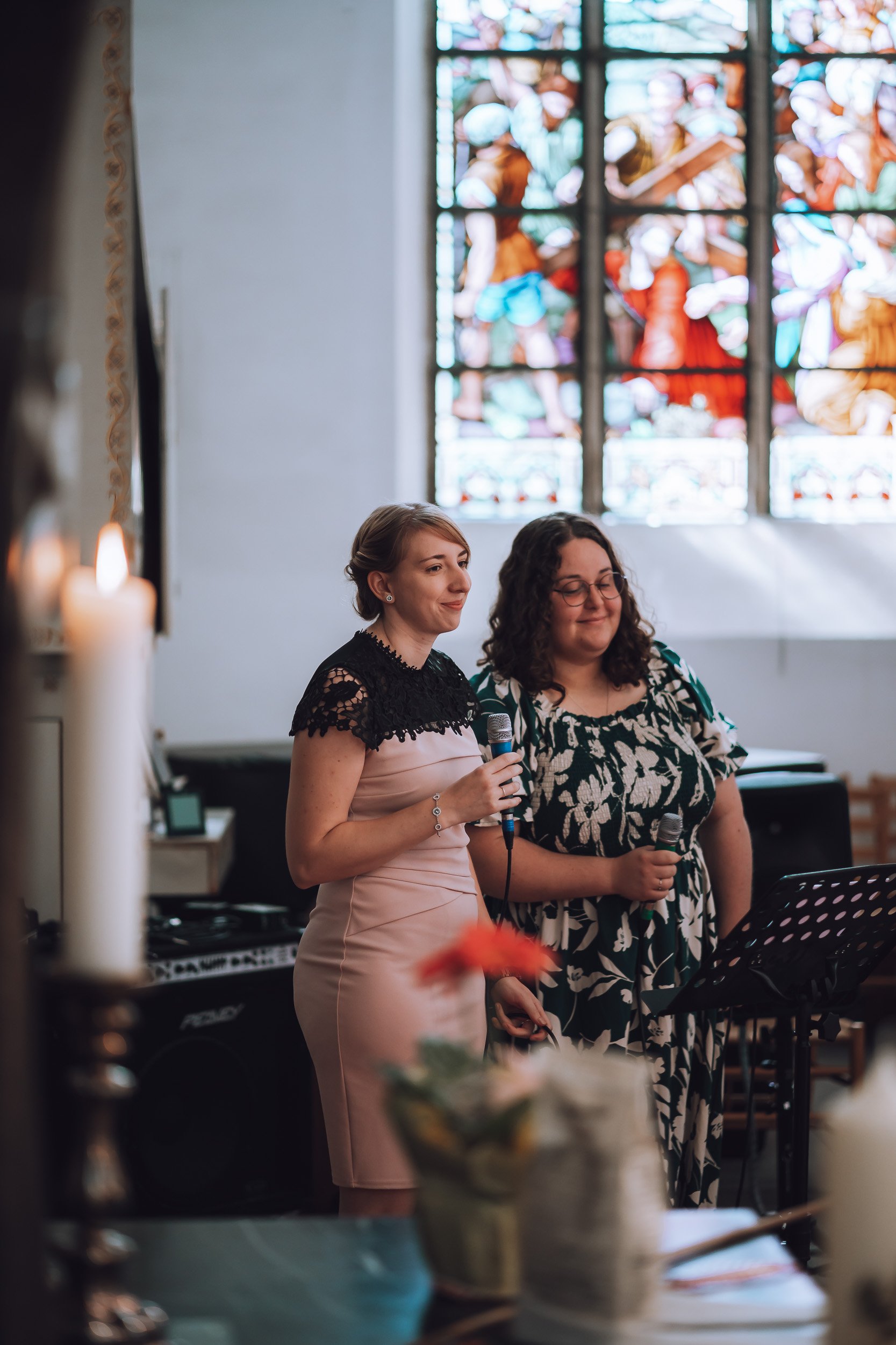 Zwei Frauen mit Mikrofonen in einer Kirche, im Hintergrund ein großes buntes Kirchenfenster.