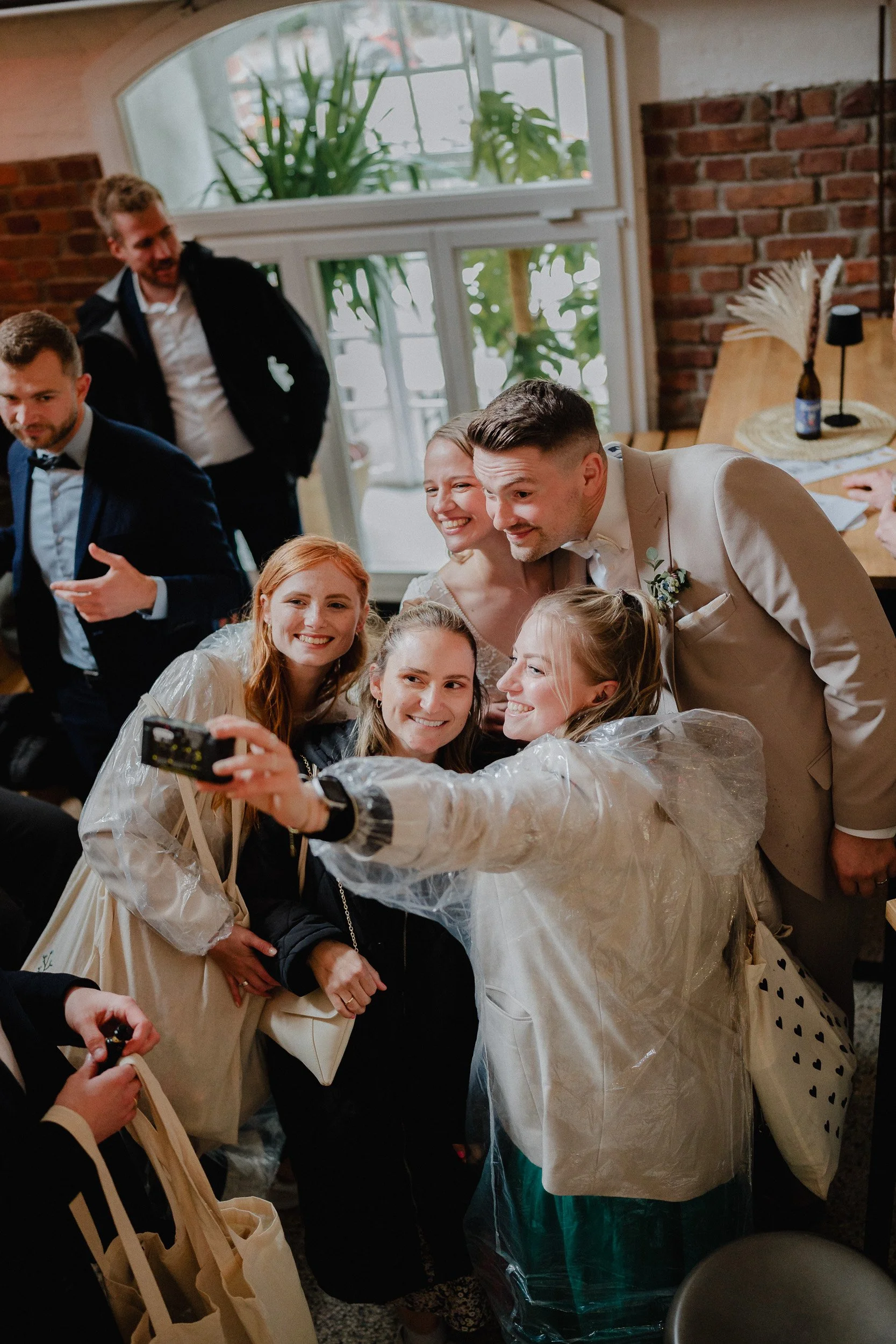Gruppe von Menschen, die für ein Selfie posieren, bei einer Hochzeitsfeier in einem gemütlichen Raum mit großen Fenstern und Pflanzen im Hintergrund.