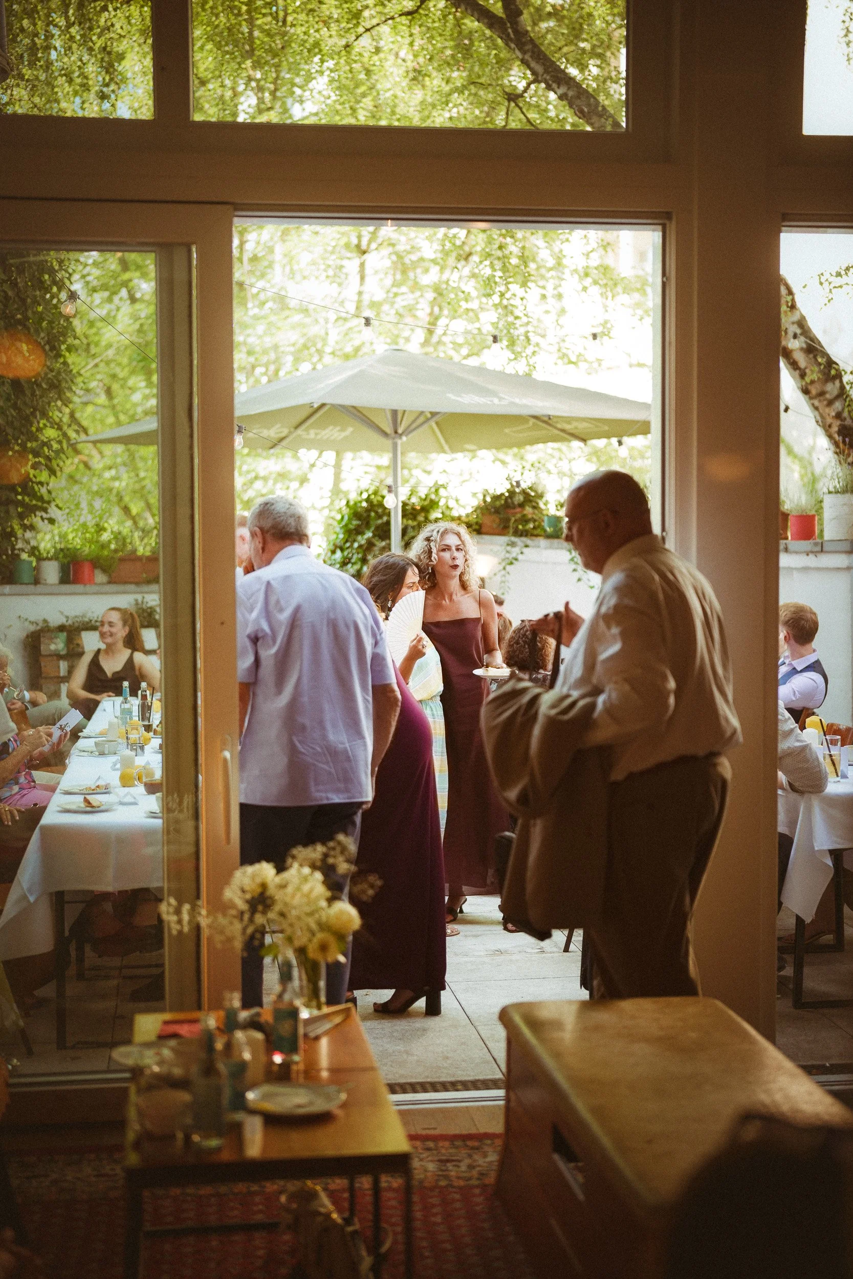Menschen feiern draußen bei einem Essen, einige stehen, andere sitzen, im Hintergrund ein Sonnenschirm, draußen grüne Bäume, im Vordergrund Blumen auf einem Tisch.