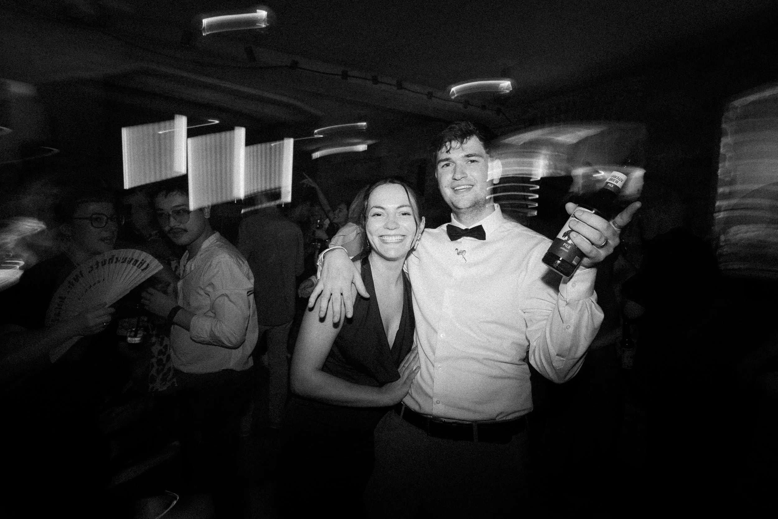 Zwei lachende junge Erwachsene auf einer Party, der Mann trägt ein weißes Hemd mit Fliege und hält ein Getränk, die Frau trägt ein dunkles Kleid.