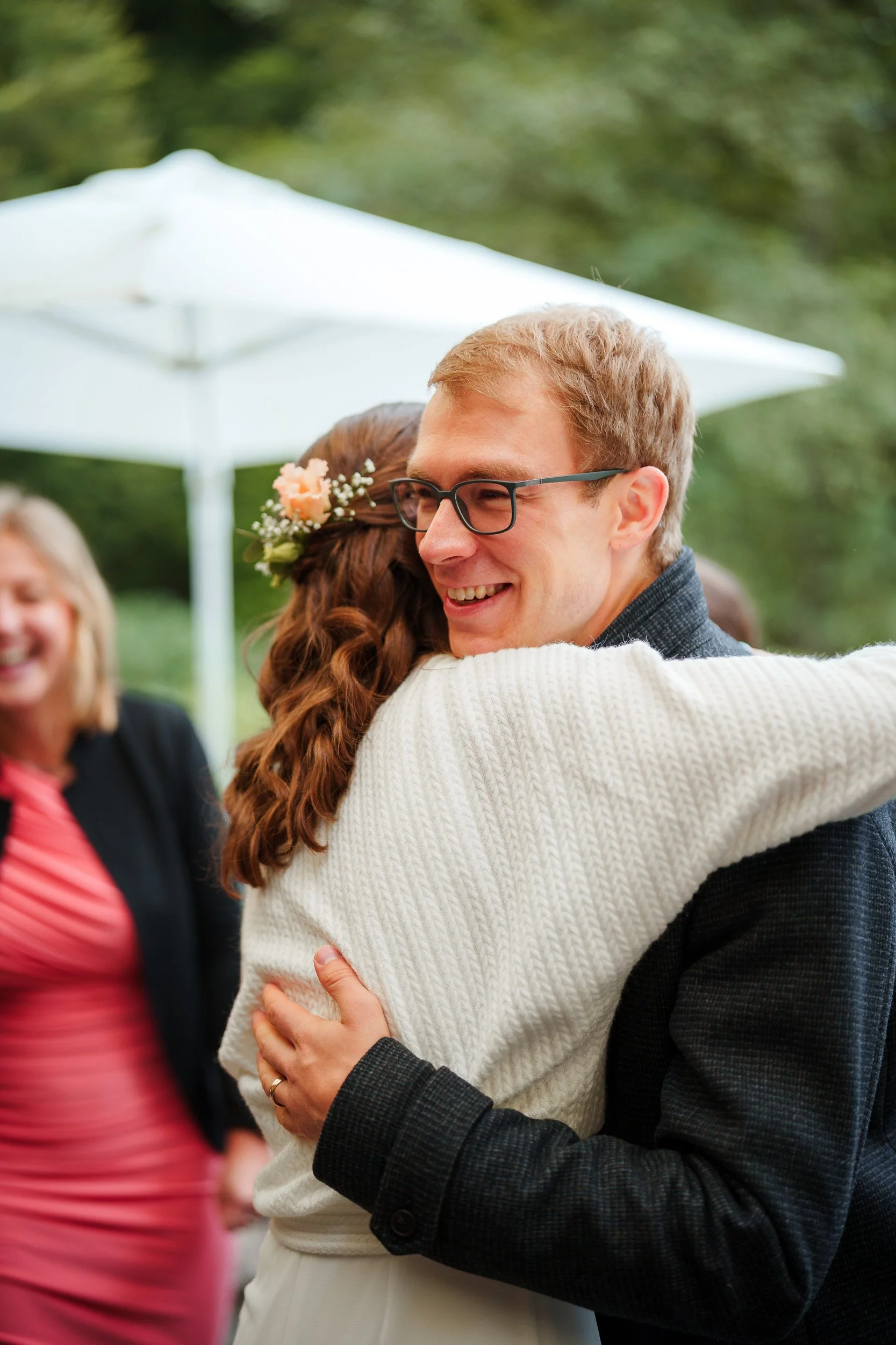 Ein Mann und eine Frau umarmen sich glücklich bei einer Feier im Freien, im Hintergrund sind andere Gäste und ein weißer Sonnenschirm zu sehen.