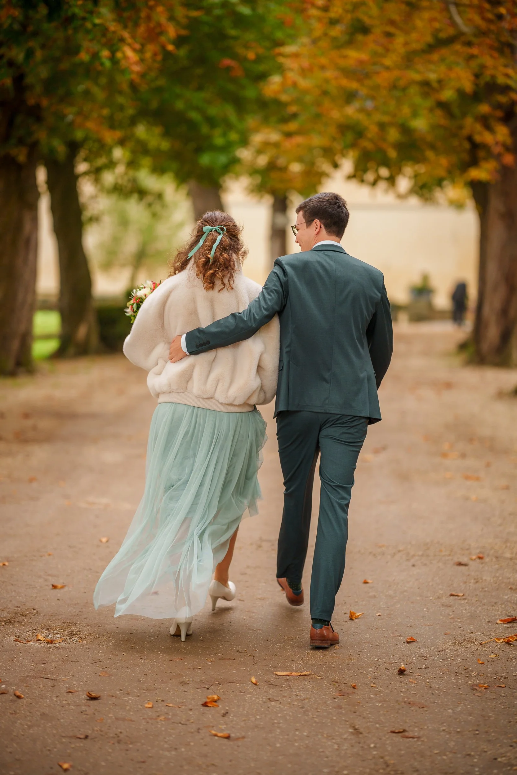 Ein Paar, vermutlich Braut und Bräutigam, läuft arm in arm durch einen Gartenweg mit herbstlichen Bäumen. Sie sind in festlicher Kleidung unterwegs, der Mann trägt einen dunklen Anzug, die Frau eine lange, fließende Tüllhose, und sie hält einen Blume