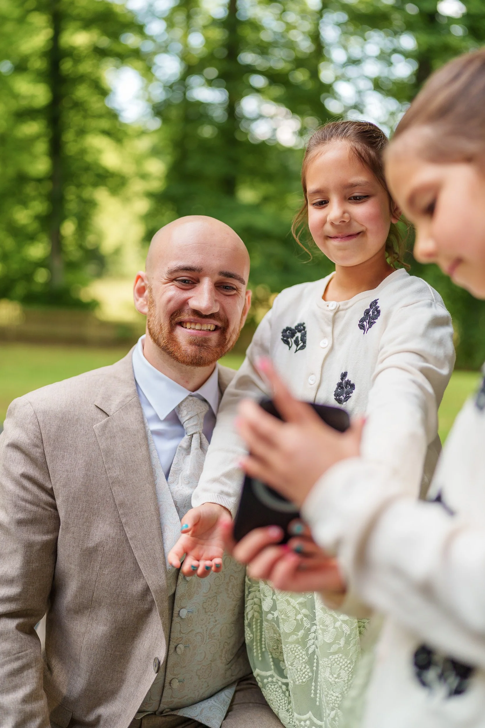 Ein Mann im Anzug mit zwei Mädchen, die draußen in einem Park sind, zeigt auf ein Smartphone, während die Mädchen lachen und sich das Gerät ansehen.