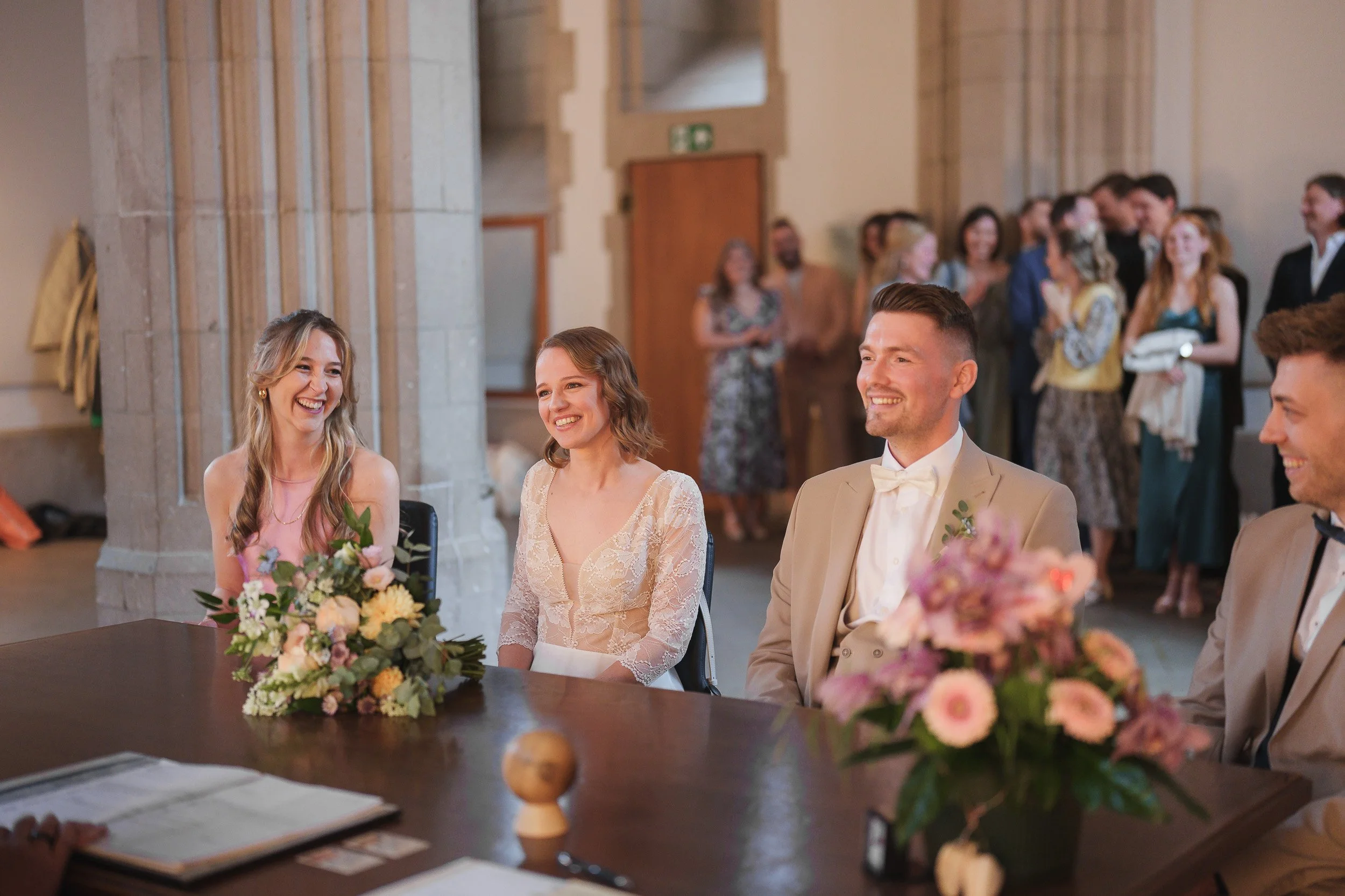 Hochzeitsszene in einer Kirche mit dem Brautpaar und Gästen, lachende Frauen sitzen am Tisch, Blumen und Dokumente auf dem Tisch, im Hintergrund weitere Gäste in Gesellschaft.