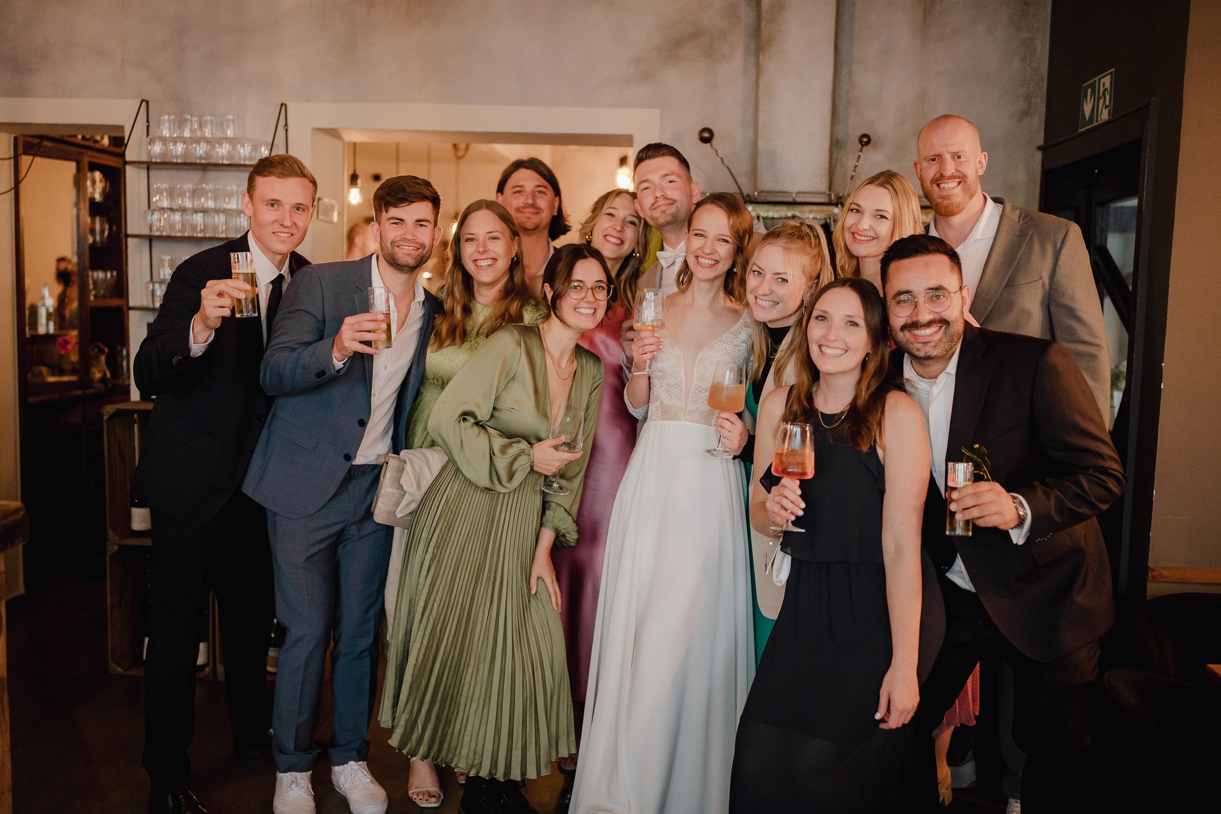Eine Gruppe von Menschen bei einer Feier, alle halten Getränke und lächeln. Es ist eine festliche Atmosphäre, vermutlich eine Hochzeit.