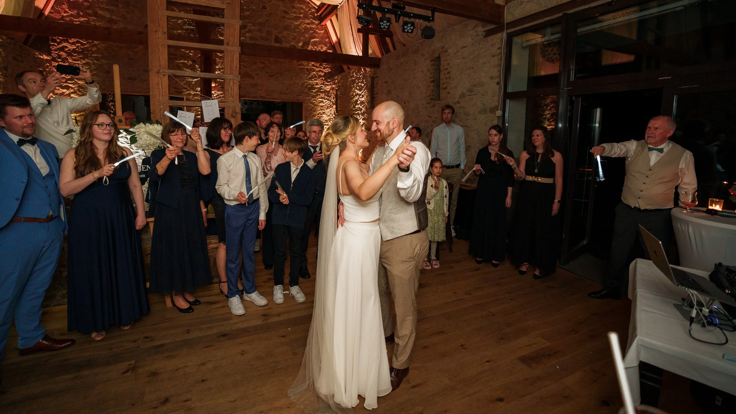 Ein Brautpaar tanzt bei einer Hochzeit, umgeben von Gästen in einem festlich dekorierten Raum mit Holzdecken. Die Gäste halten Wunderkerzen in den Händen.