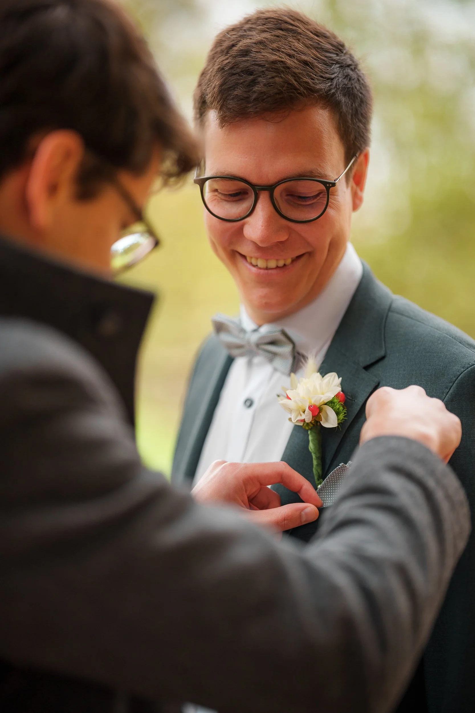 Zwei Männer, einer in einem dunklen Anzug mit Brille, der den anderen, einen Bräutigam, an einem grauen Anzug mit Fliege und Blumenanstecker befestigt, bei einer Hochzeitsfeier im Freien.