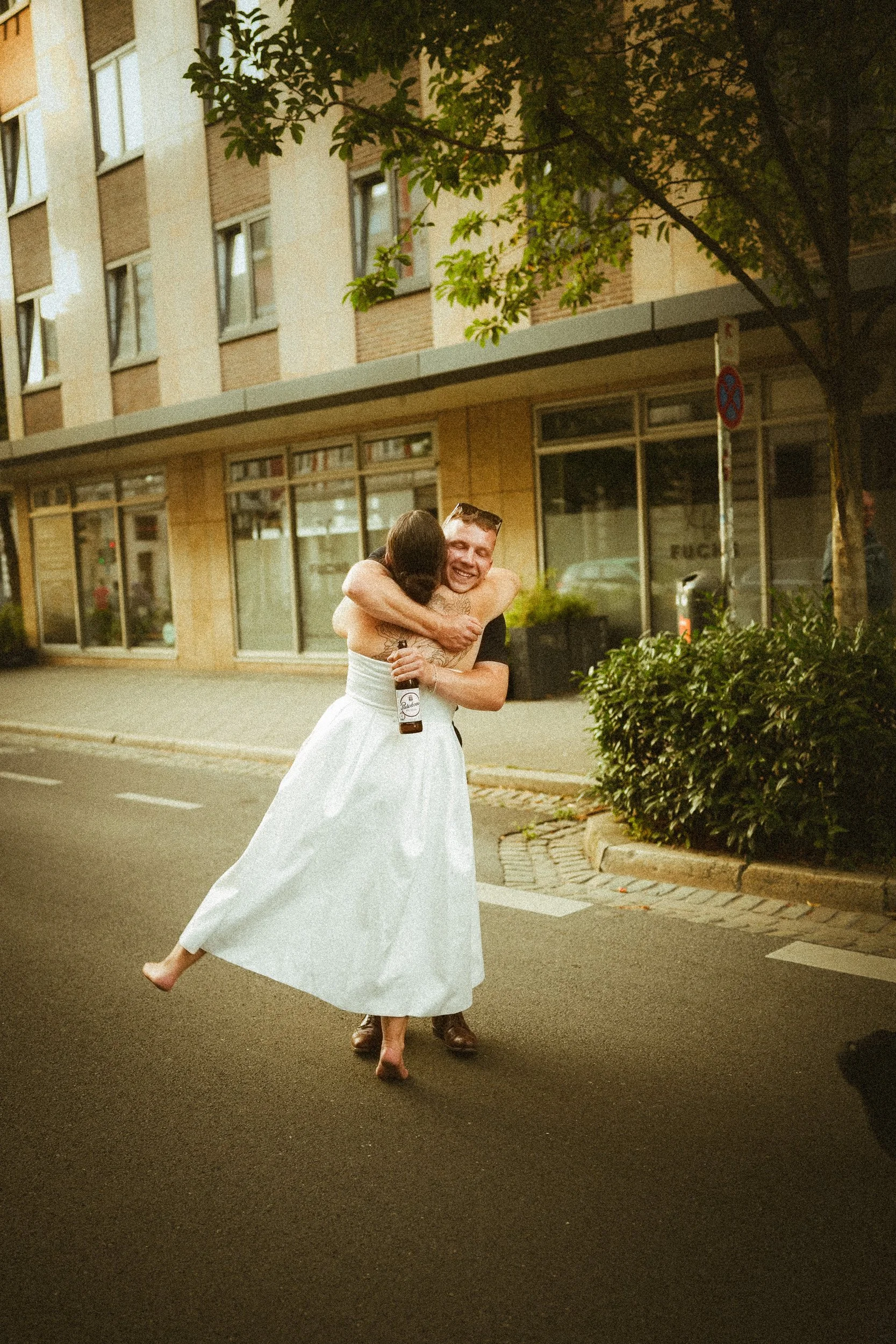 Zwei Menschen, die sich auf einer Straße umarmen, eine Person in einem weißen Kleid und die andere in dunkler Kleidung, bei Nacht, in einer urbanen Umgebung mit Gebäuden und Bäumen.