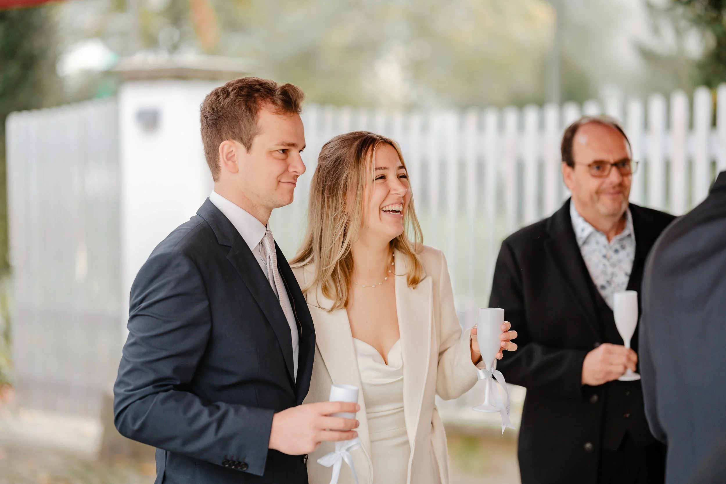 Menschen bei einer Feier, eine Frau lacht und hält ein Glas Champagner, zwei Männer im Anzug, im Hintergrund eine weiße Zaun