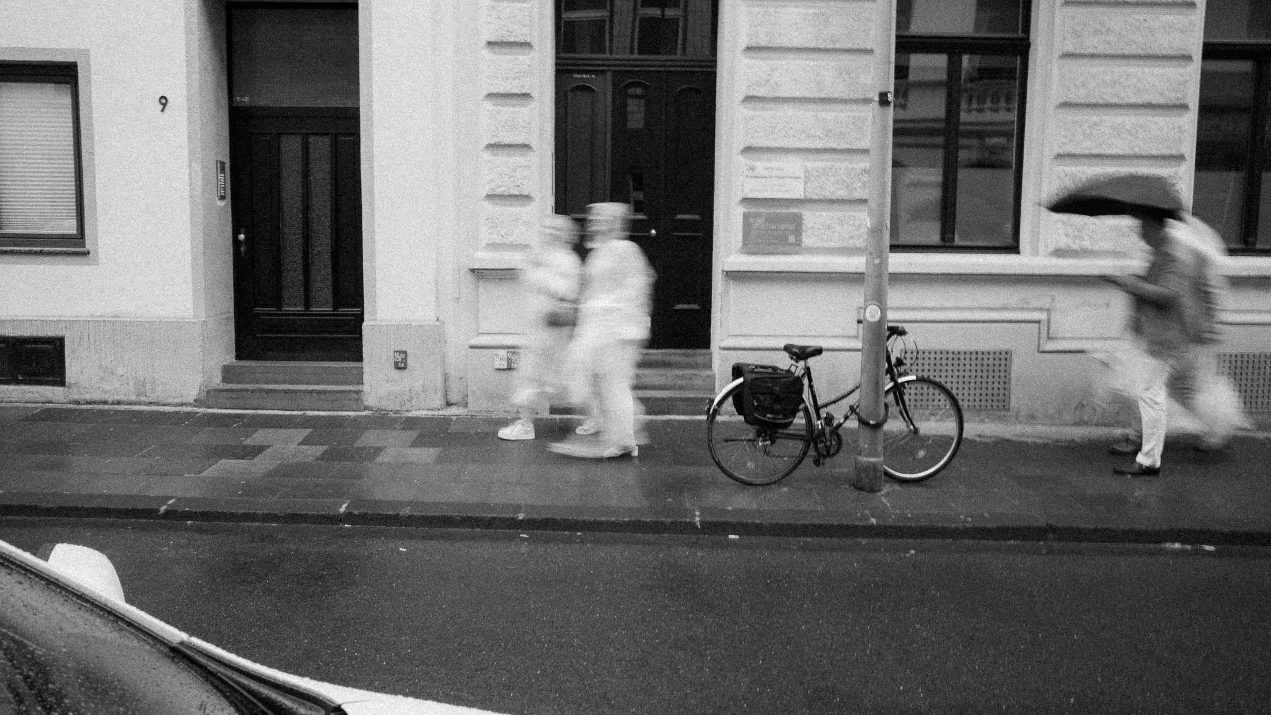 Straßenansicht mit mehreren Menschen, die im Regen gehen, eine Person hält einen Regenschirm, ein Fahrrad steht an einem Laternenpfahl, alles in Schwarzweiß aufgenommen.