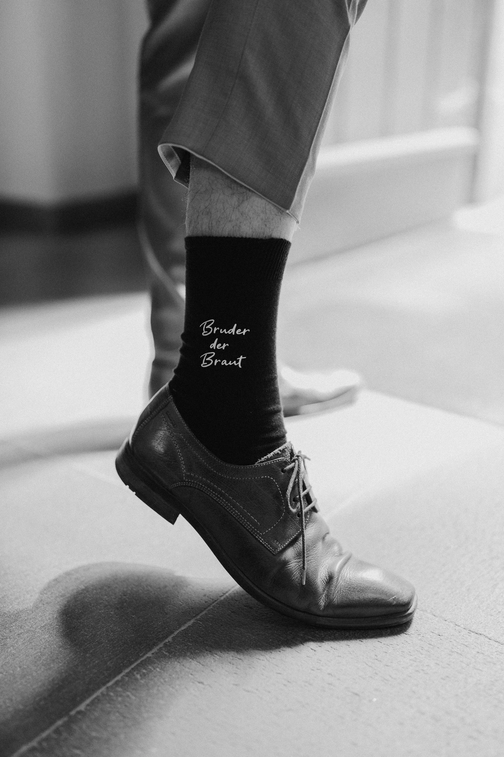 Schwarze Socke mit weißen Schriftzug 'Bruder der Braut' auf einem braunen Schuh, der auf einem Boden steht, in Schwarz-Weiß-Fotografie.