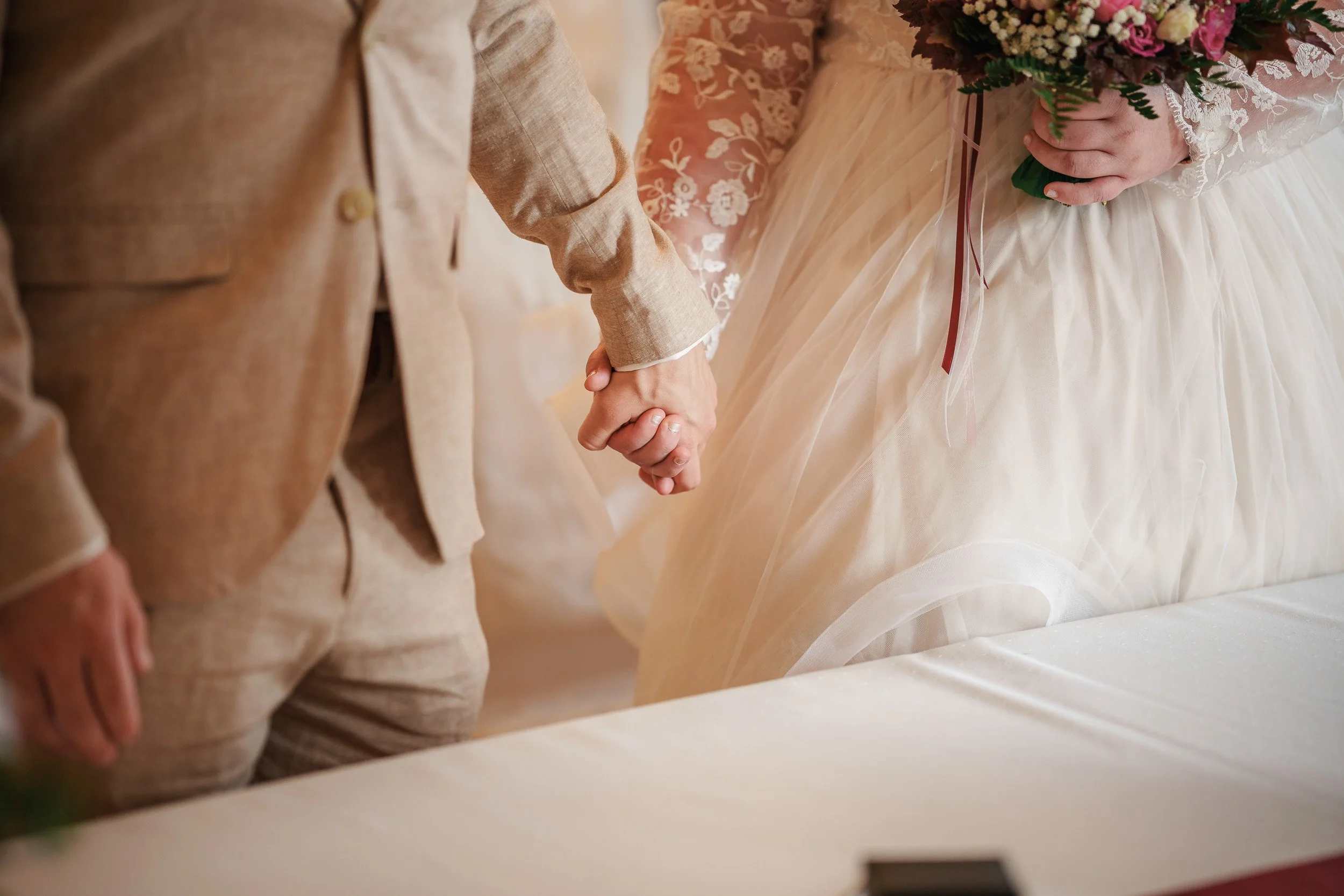 Ein Brautpaar hält sich bei einer Hochzeitszeremonie an den Händen. Die Braut trägt ein weißes Kleid mit Spitze und einen Brautstrauß, während der Bräutigam einen hellbraunen Anzug trägt.