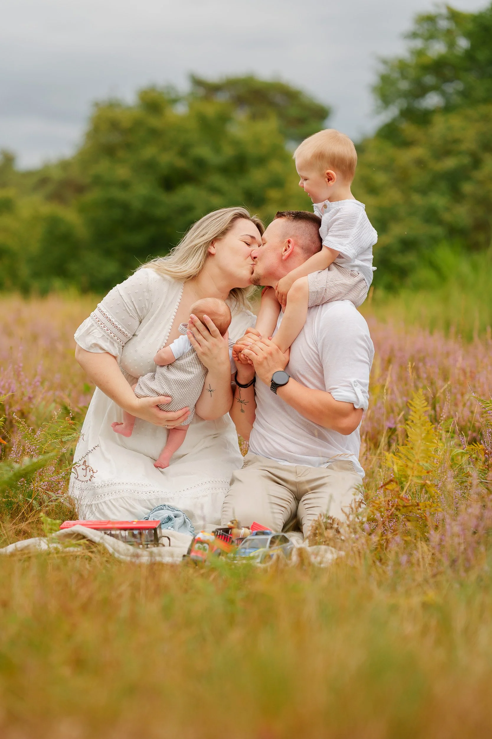 Familie bestehend aus Mutter, Vater, einem kleinen Jungen und einem Neugeborenen bei einem Kuss im Freien, umgeben von Blumen und Bäumen.