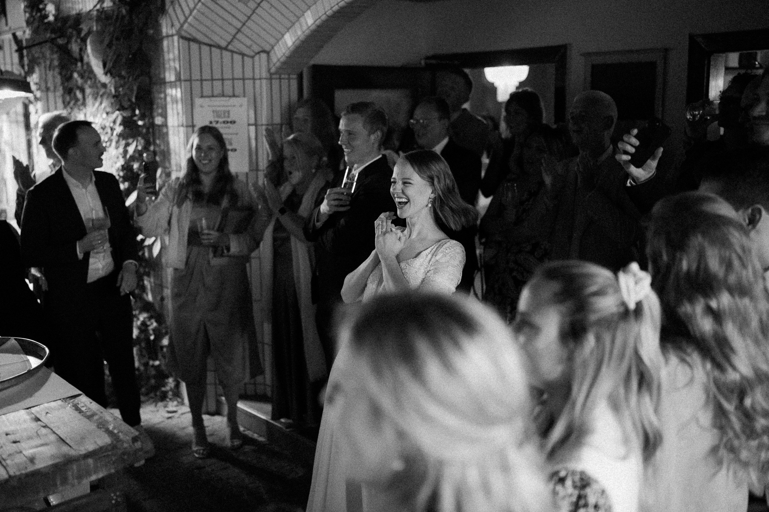 Menschen auf einer Feier, einige lachen, andere fotografieren, im Hintergrund ist ein dunkler Raum, wohl auf einer Party oder Hochzeit.