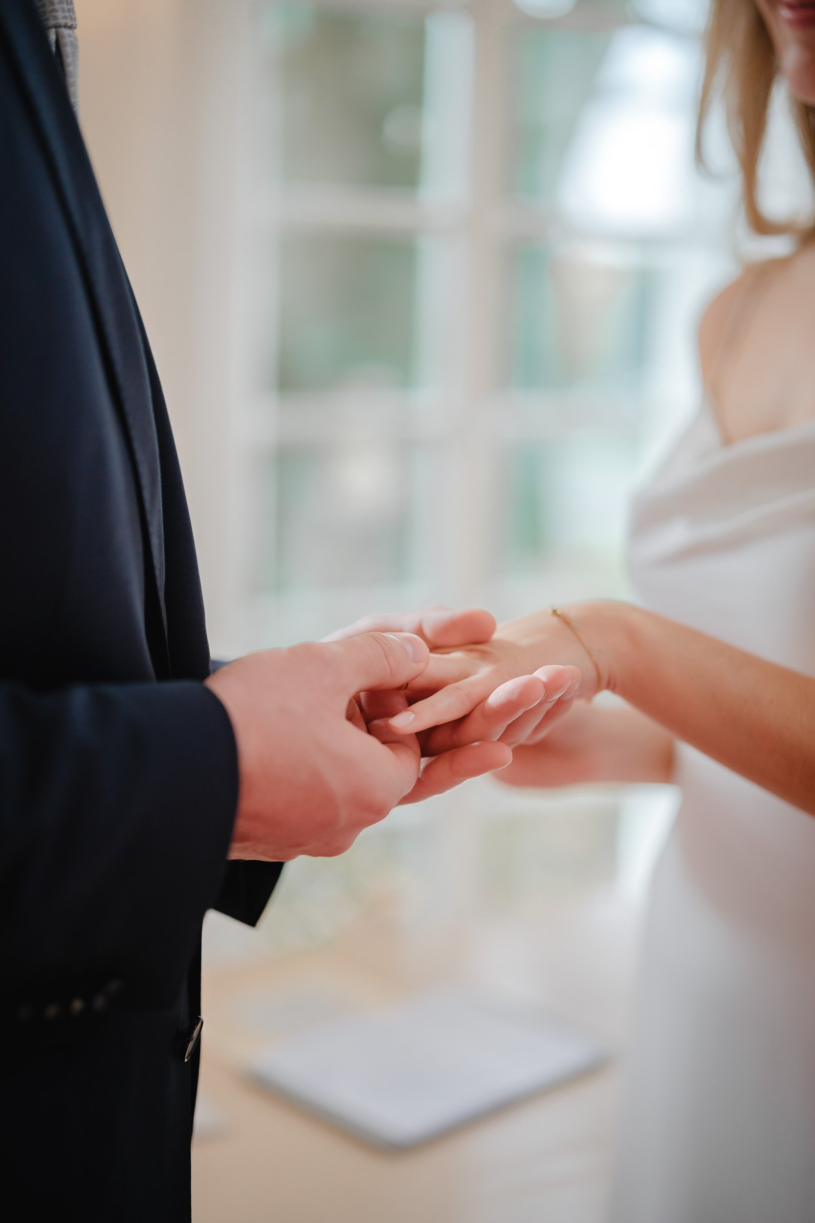 Ein Mann in einem schwarzen Anzug hält die Hand einer Frau in einem Hochzeitskleid beim Austausch der Ringe in einem Raum mit großen Fenstern.