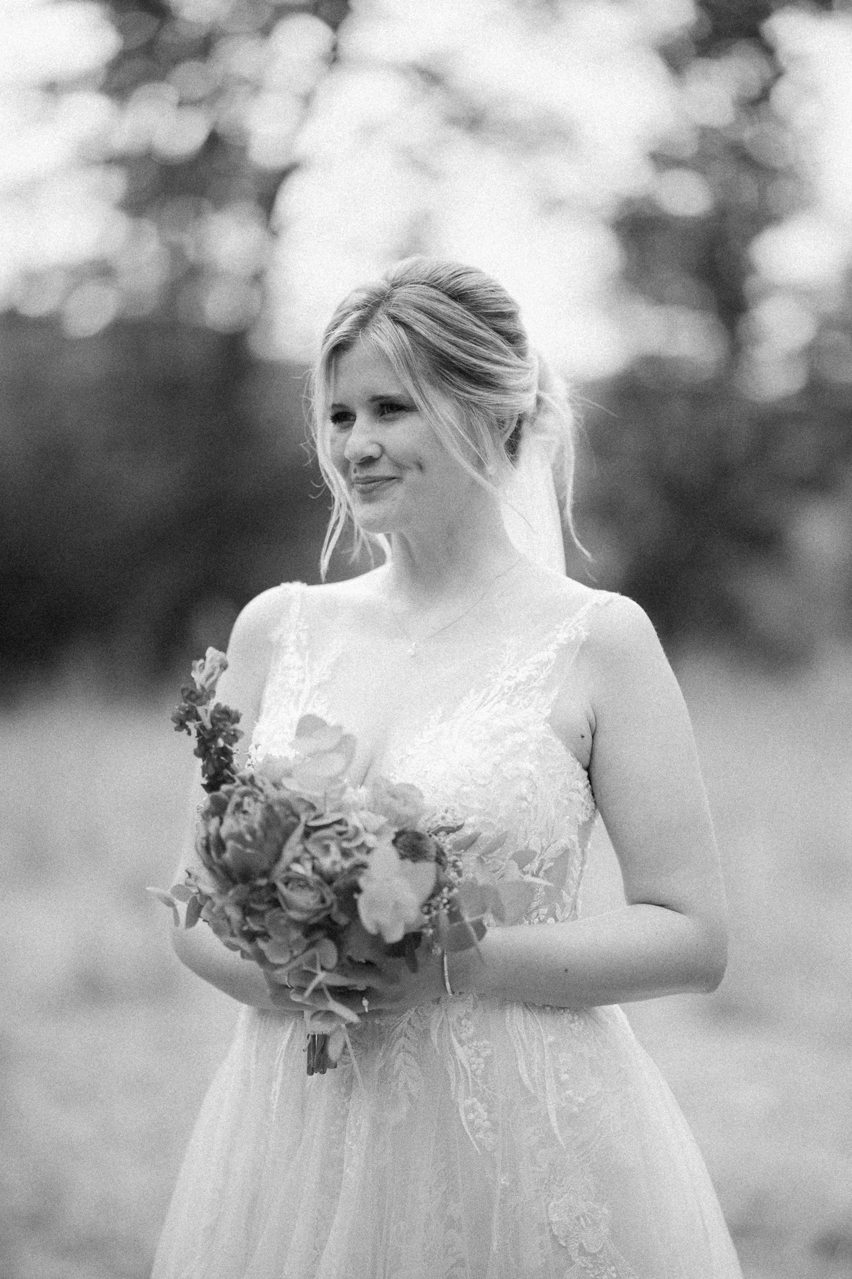 Braut in Hochzeitskleid hält Blumenstrauß in den Händen, lächelt im Freien, unscharfer Hintergrund.
