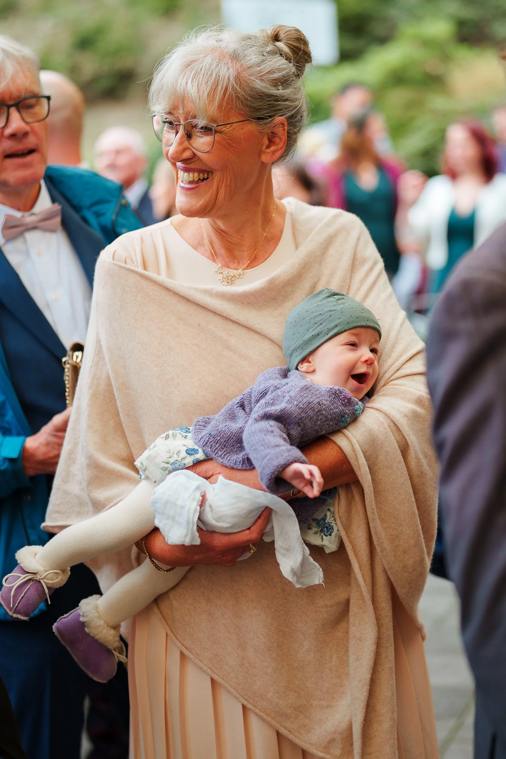 Eine ältere Frau hält ein lachendes Baby in den Armen bei einer gesellschaftlichen Veranstaltung im Freien, umgeben von mehreren Menschen.