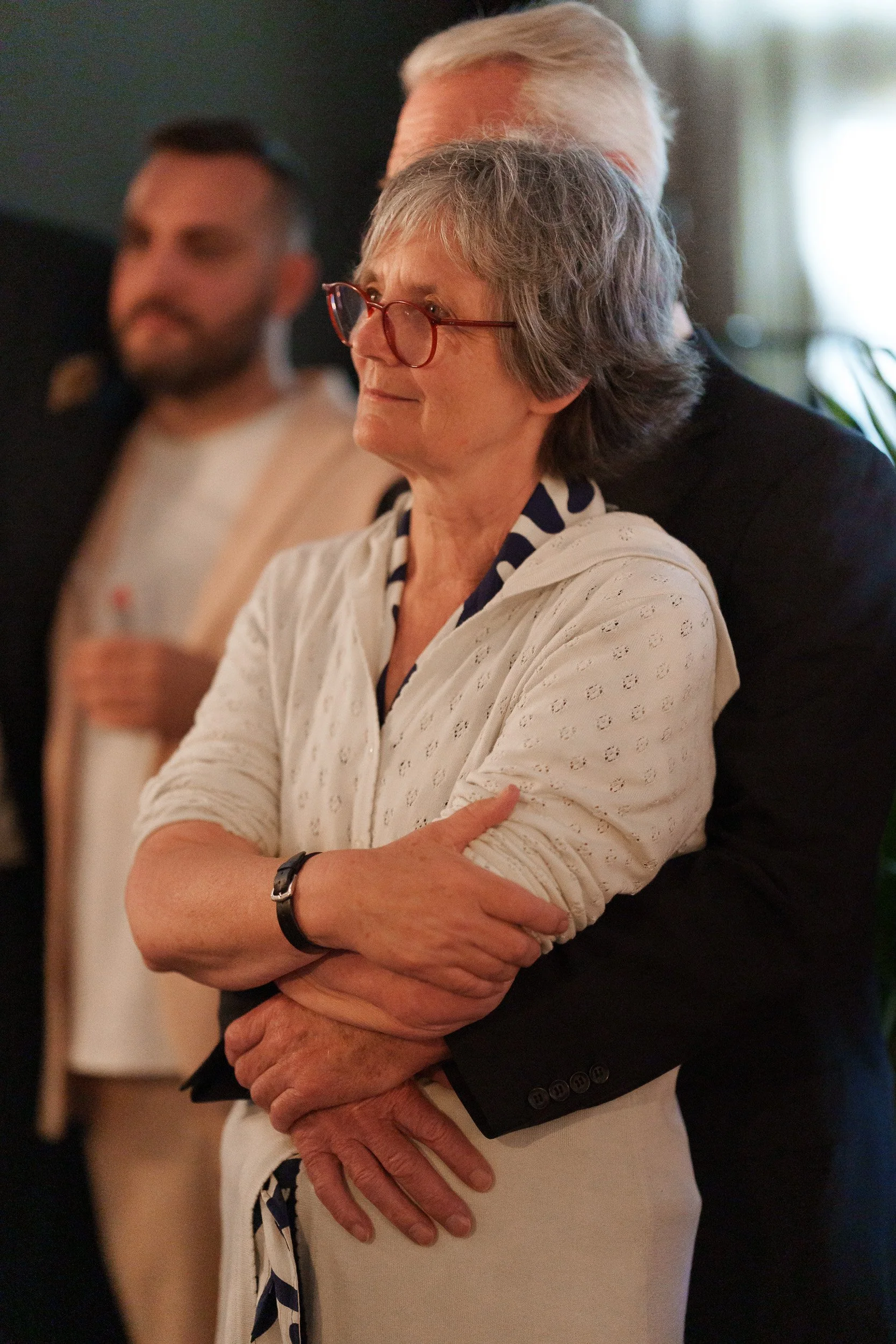 Ältere Frau mit Brille, trägt eine weiße, perforierte Jacke und steht vor einem Mann, der sie umarmt. Im Hintergrund sind weitere Personen sichtbar, die bei einer Zusammenkunft oder Veranstaltung anwesend sind.