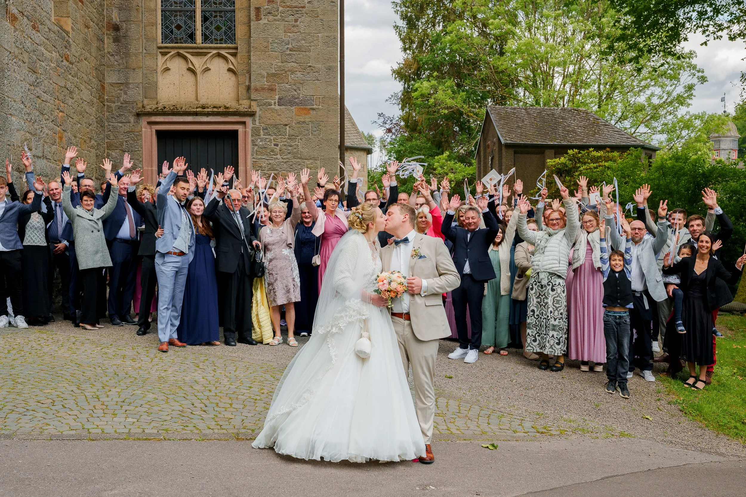 Ein Brautpaar küsst sich vor einer Gruppe von Gästen bei einer Hochzeitsfeier im Freien vor einer alten Steinmauer mit Bäumen im Hintergrund.