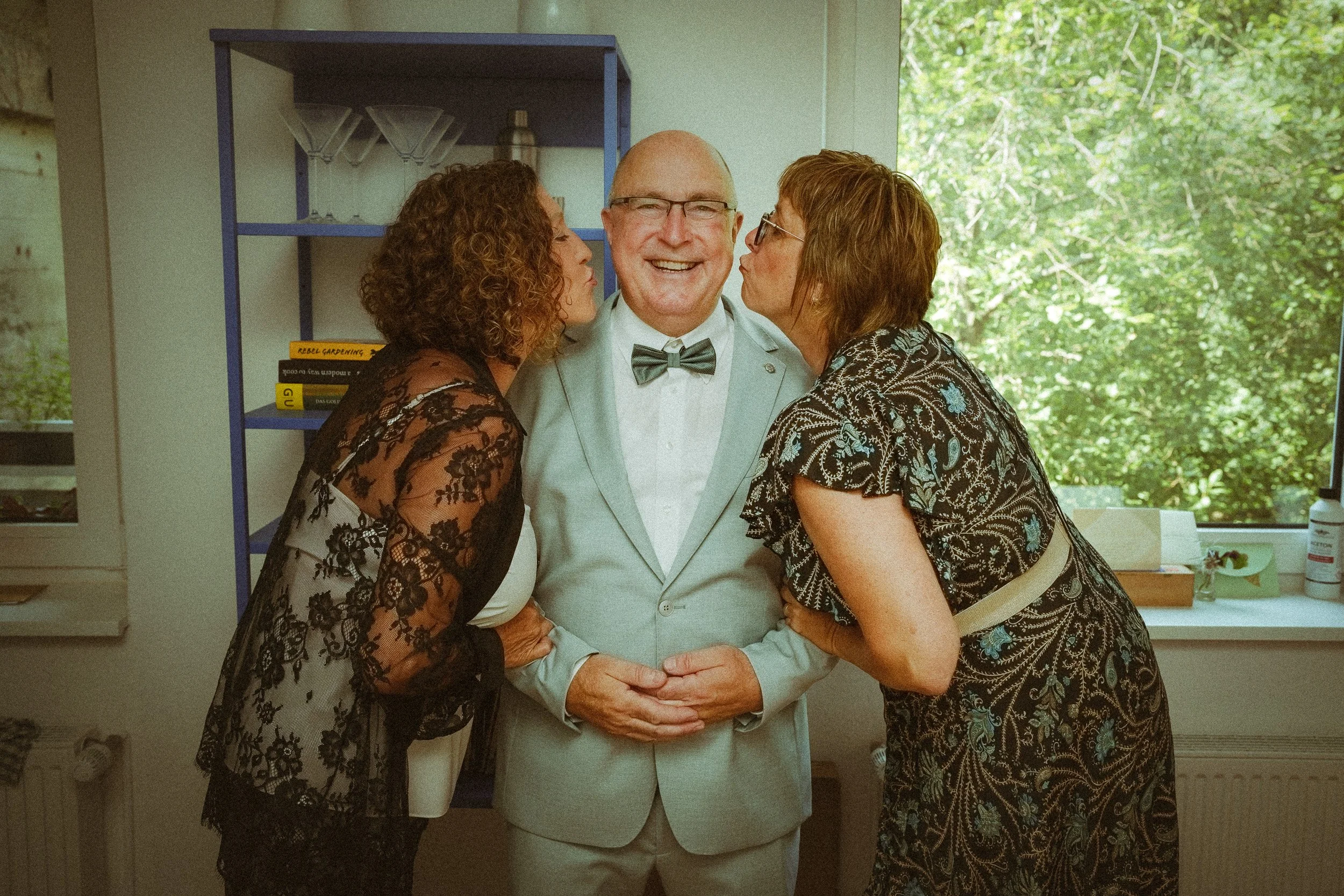 Ein Mann in einem weißen Anzug und einer Fliege wird von zwei Frauen geküsst, die an seinen Wangen küssen, während er lächelnd in die Kamera schaut. Sie stehen in einem Raum mit einem Fenster, durch das Bäume zu sehen sind.