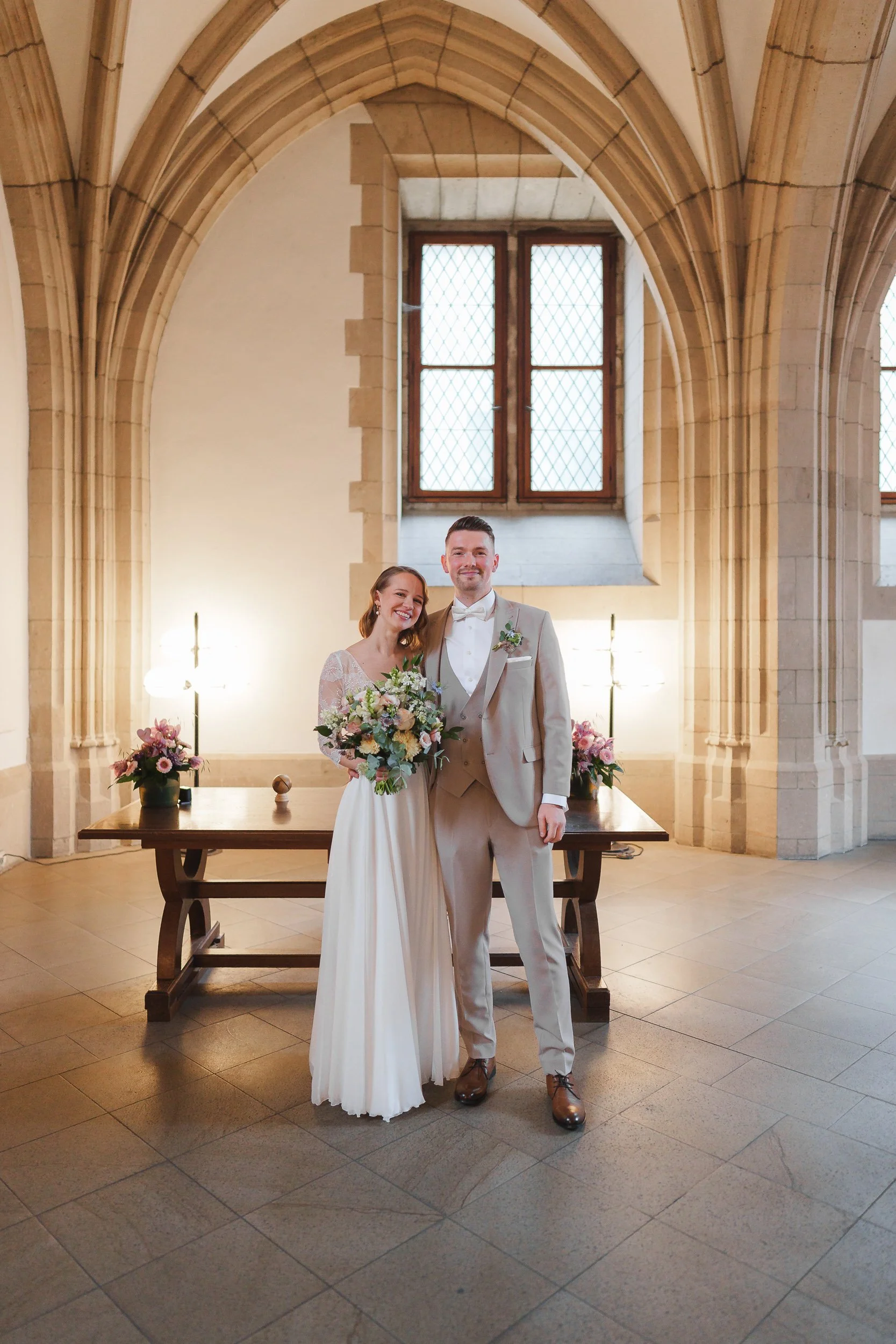 Ein Brautpaar steht in einer alten Kirche, lächelnd und im Mittelpunkt. Die Braut trägt ein langes weißes Kleid und hält einen Blumenstrauß, der Bräutigam trägt einen hellen Anzug mit Fliege.