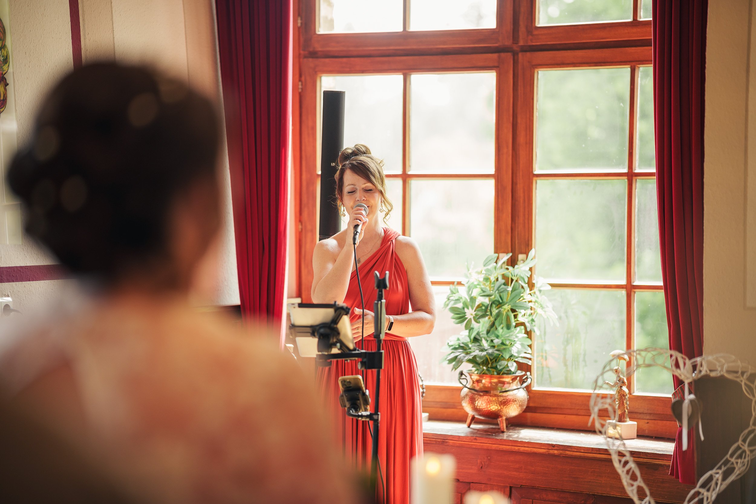 Eine Frau in rotem Kleid singt mit einem Mikrofon vor einem Holzfenster, während eine Person im Vordergrund sie anschaut.