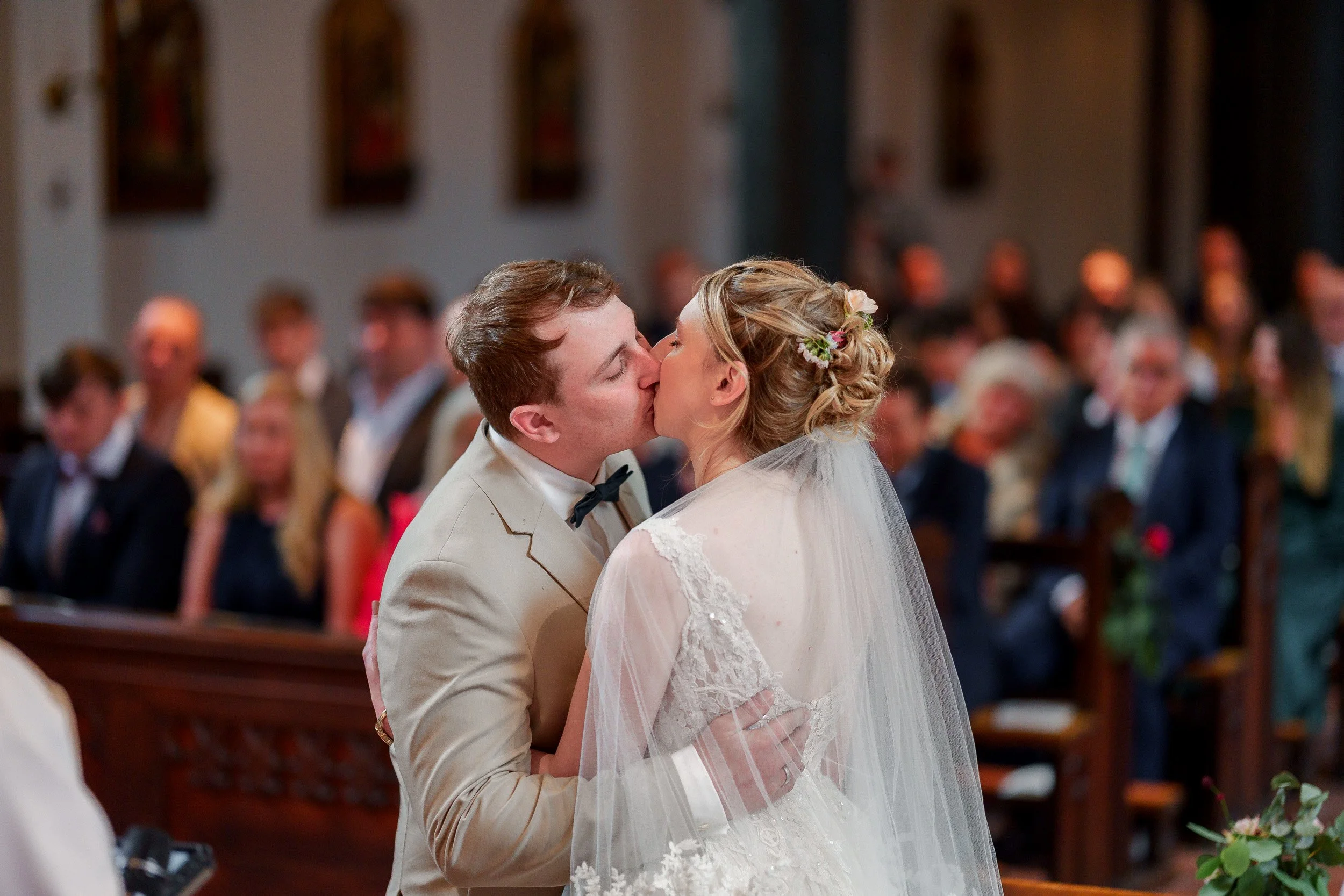 Ein Brautpaar küsst sich während einer Trauung in einer Kirche, im Hintergrund sitzen Gäste.