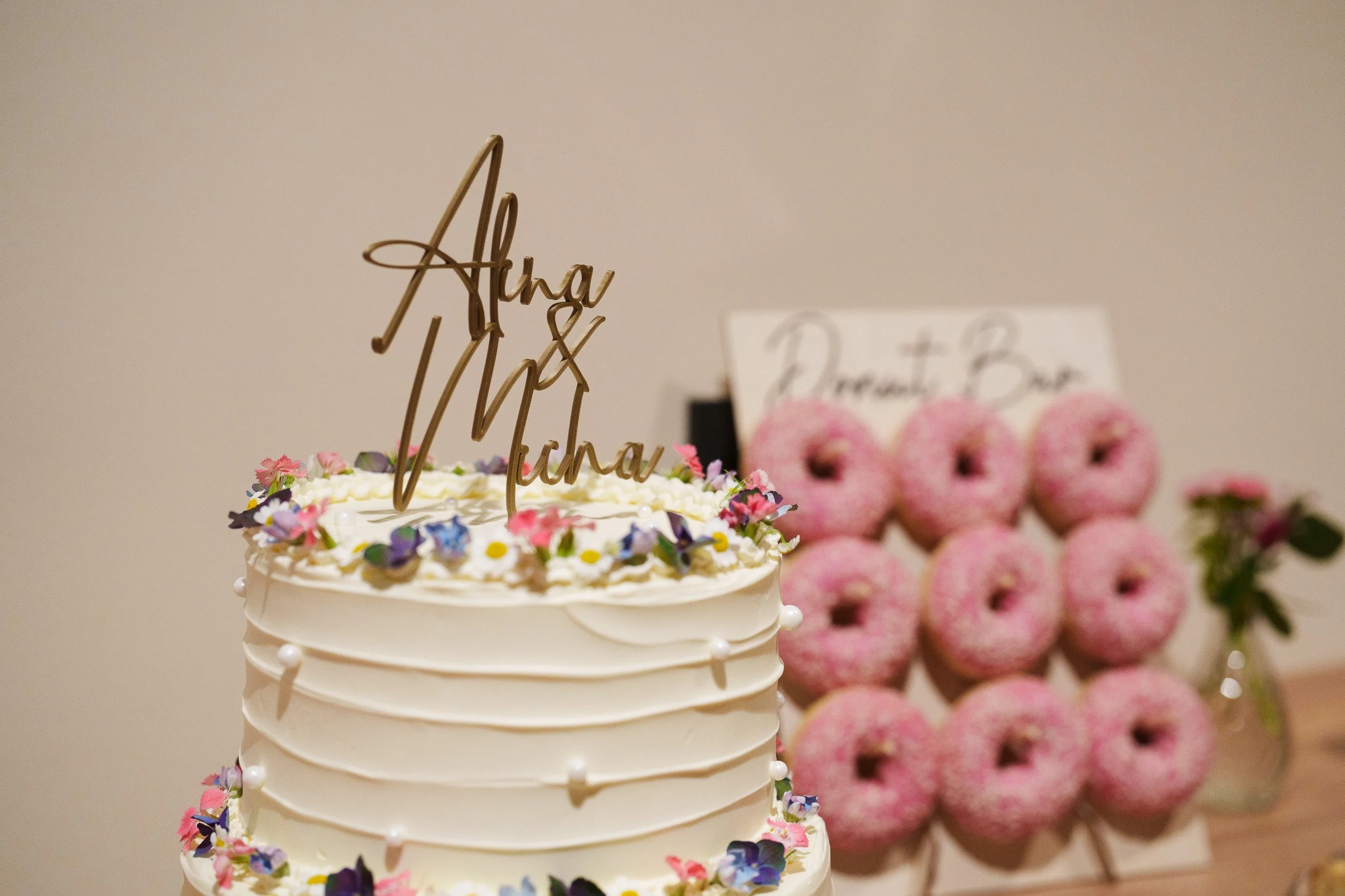 Ein weißer Hochzeitstorte mit Blumen und einem goldenen Namensschild, im Hintergrund eine Schale mit rosa Donuts }