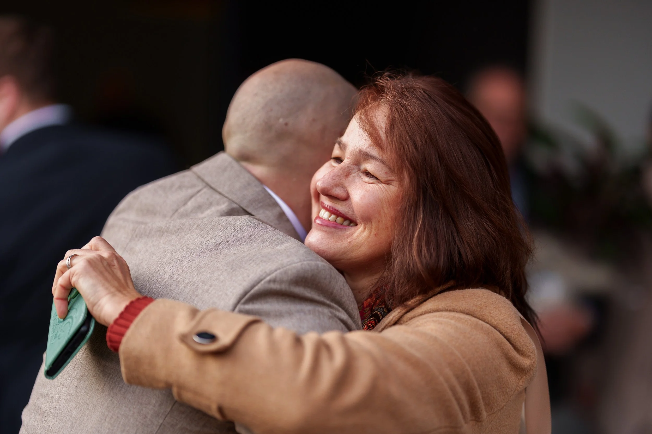 Zwei Menschen umarmen sich herzlich und lächeln. Im Hintergrund sind weitere Personen sichtbar.