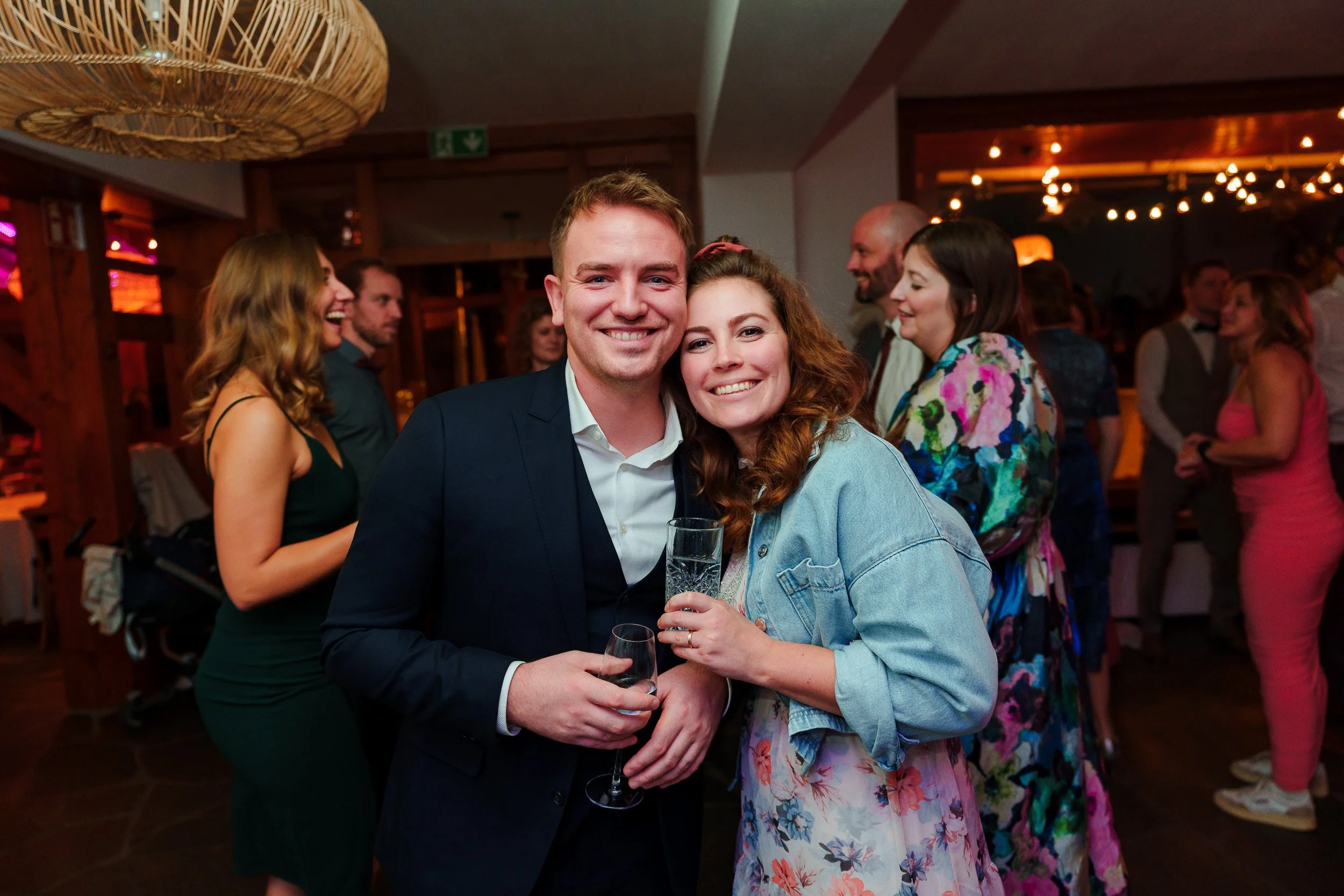 Ein lachendes Paar bei einer Feier, das sich umarmt, mit Gläsern in der Hand, im Hintergrund andere Gäste in einer festlichen Stimmung in einem dunkel beleuchteten Raum.
